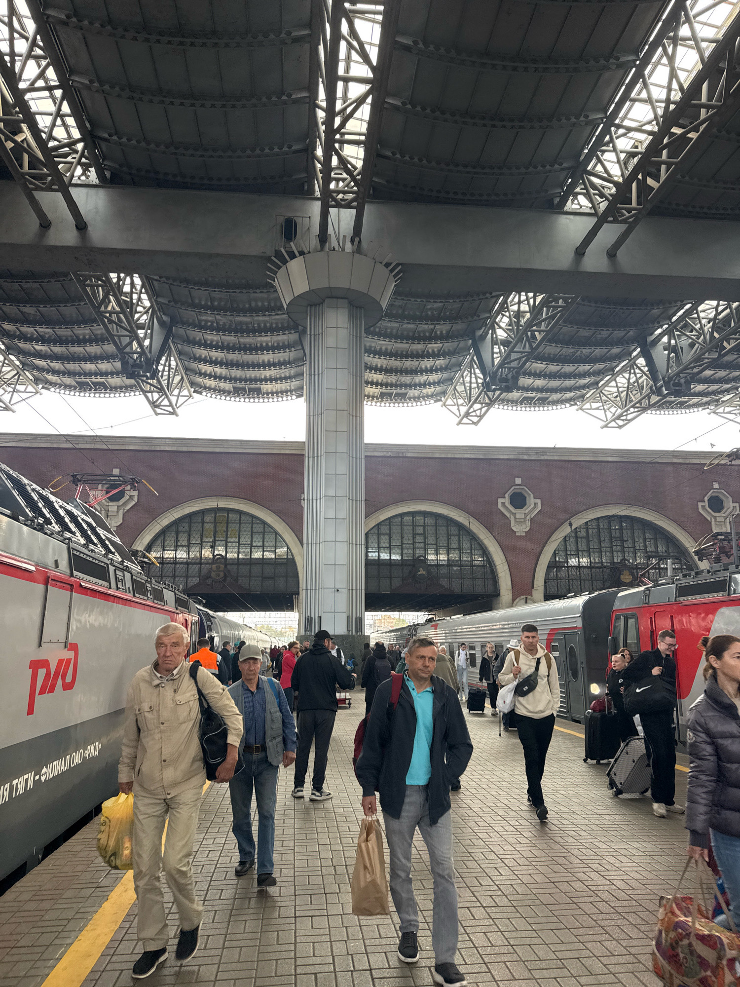 The image depicts a busy train station platform in Moscow, Russia. A red and silver train is parked on the tracks to the left, bearing the Russian Railways logo. Passengers are seen walking along the platform, some carrying luggage. The station features a large, arched roof structure supported by columns, typical of many historic train stations. The architecture suggests a substantial, possibly older, building. The lighting indicates it is daytime, with overcast conditions. The people are dressed in casual, everyday clothing, suggesting a normal day of travel. The overall scene conveys the hustle and bustle of a major transportation hub.