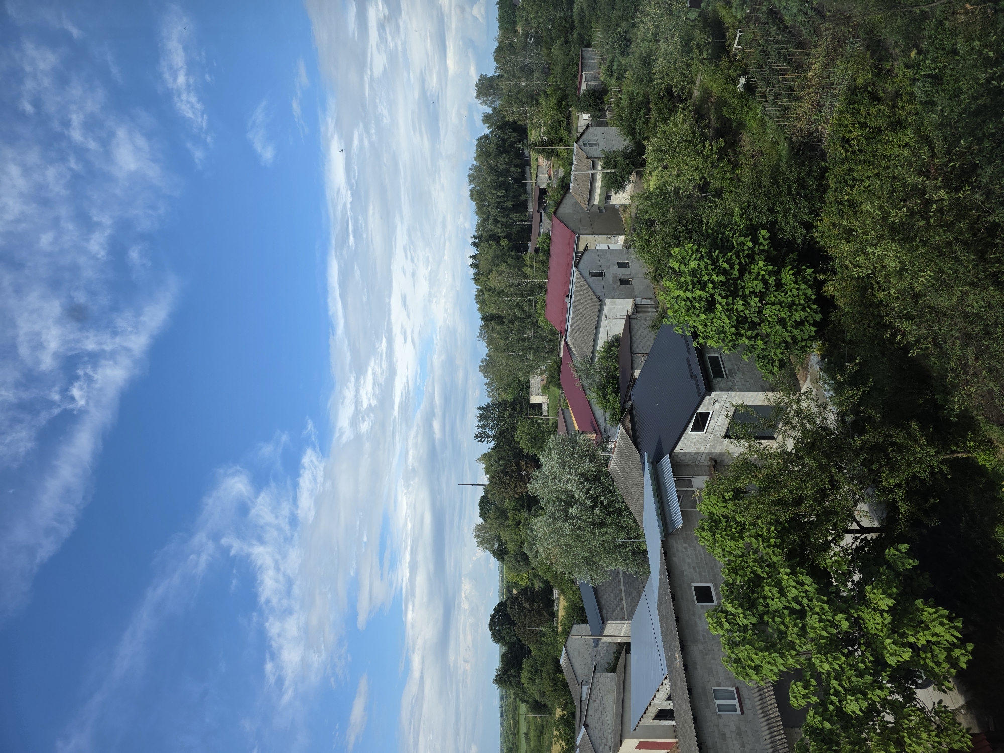 This is a daytime, exterior, high-angle, medium shot of a residential area in Dubno, Ukraine. The scene is dominated by a bright blue sky with scattered white clouds. In the foreground, several low-rise buildings with grey concrete block walls and various roof types (some corrugated metal, some dark asphalt shingles, and one with red shingles) are visible. Some of these buildings appear to be garages or workshops. Lush green trees and foliage surround and are interspersed among the buildings. Beyond the immediate cluster of houses, a dense line of trees forms a backdrop. In the distance, a flat, green agricultural field can be seen under the expansive sky. There are no people or animals visible in the image. The overall impression is of a rural or suburban residential area.