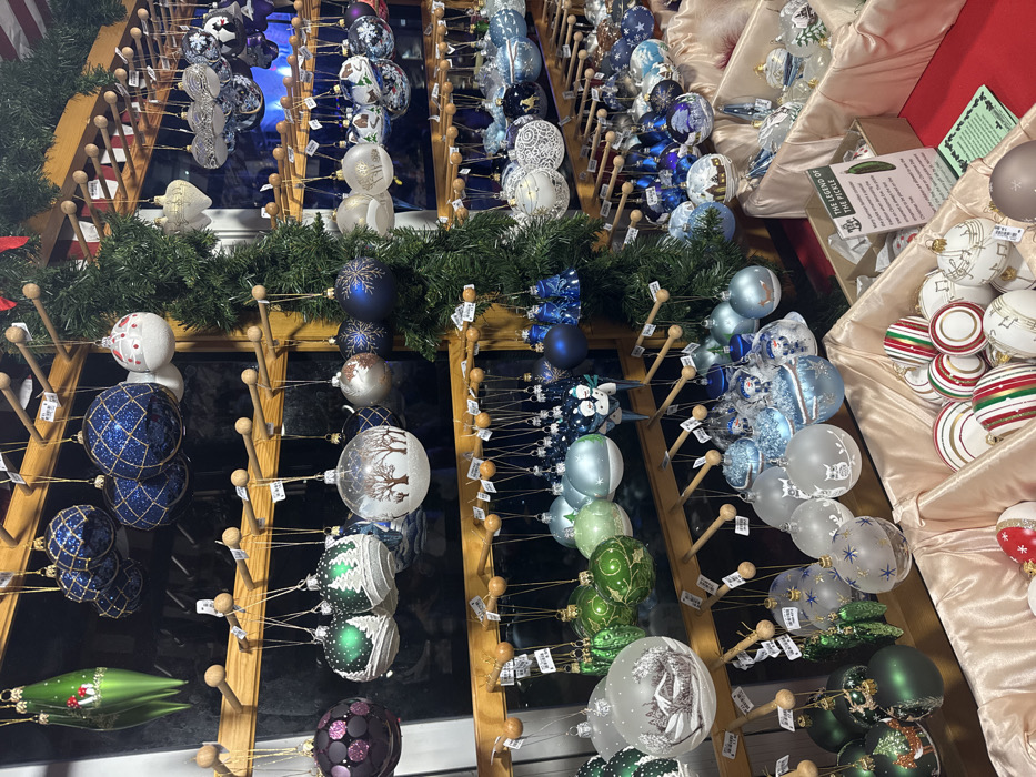 A display of Christmas ornaments features various colors and designs. Blue, white, green, and purple ornaments hang on wooden racks adorned with evergreen garlands. Some ornaments have intricate patterns, while others feature winter scenes. Additional ornaments in white, gold, red, and green fill beige-lined shelves. Small price tags dangle from the ornaments.