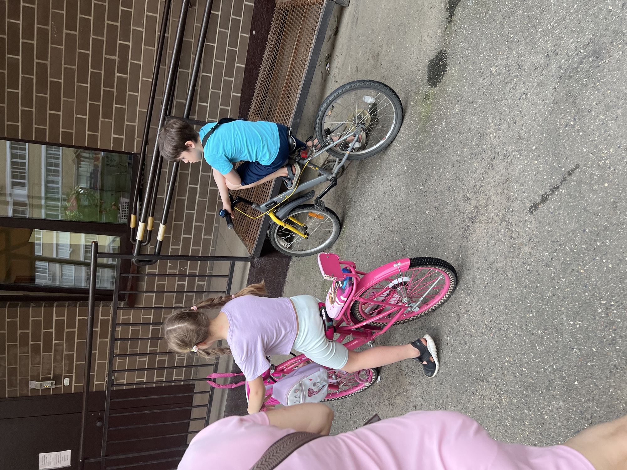 The image displays an outdoor scene in Krasnodar, Russia, featuring two children on bicycles and a partially visible adult, set against the backdrop of a brick building.
A young girl, with light brown hair styled in two pigtails, is seated on a bright pink bicycle, facing away from the camera and slightly to the left. She wears a light purple short-sleeved crop top, light-colored cycling shorts, and dark sandals with white soles. Her bicycle is equipped with a coiled pink chain lock on the handlebars, a small light purple backpack hanging from the handlebars, and a patterned pink seat cover with an additional small pink seat/rack over the rear fender.
To the right of the girl, a young boy with short brown hair is seated on a grey and yellow bicycle, facing left. He wears a bright blue short-sleeved t-shirt, dark blue shorts, and sandals. A dark strap, possibly from a bag, crosses his chest. His bicycle has a small blue bell on the handlebars.
On the far left, a person, likely an adult, is partially visible, showing their left side from the shoulder to the upper thigh. They are dressed in a light pink short-sleeved top and light pink bottoms, with a brown strap visible on their left shoulder.
In the background, a multi-story building constructed with brown bricks is prominent. A dark brown door is visible on the left, with an intercom system mounted on the brick wall beside it. A large, dark-framed window reflects the facade of another building featuring multiple windows and a lighter color. Below the window and spanning the mid-ground is a dark metal railing system, which includes a metal grate ramp leading up to the building entrance. Additional horizontal bars with light-colored caps are integrated into the railing structure.
The ground is a dark grey asphalt or paved surface, characterized by scattered lighter aggregate particles and several dark, irregular stains, particularly noticeable in the lower right portion of the image.