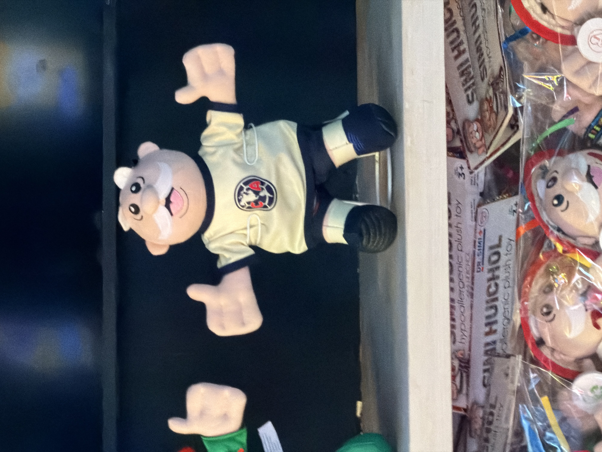The image shows a shelf with a Simi Huichol plush toy dressed in a yellow soccer uniform with the Club America logo. It has a mustache and black shoes. Other Simi Huichol plush toys are on the shelf, wrapped in plastic. The location is likely in Los Angeles, USA, due to the presence of the toys, which are often sold in Mexican pharmacies.
