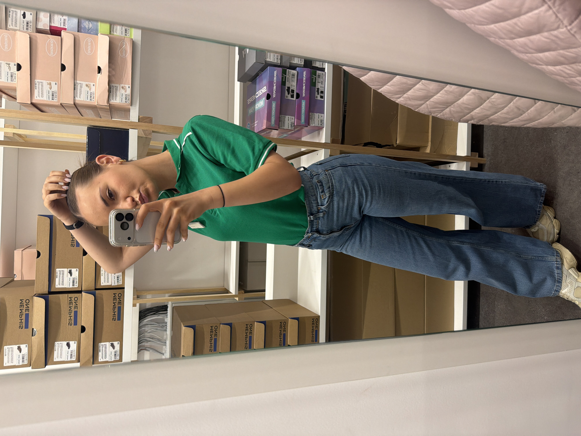 A woman is taking a selfie in a mirror inside a retail store, presumably in Sofia, Bulgaria, given the visible Cyrillic script on the shoeboxes. She is wearing a green polo shirt and blue jeans and holding a phone with her right hand. The retail environment is evident from the stacked shoeboxes lining the shelves behind her, featuring brands like Skechers and Memphis One. The lighting is bright, suggesting indoor illumination. The store appears organized, with neat rows of footwear packaging and other products.