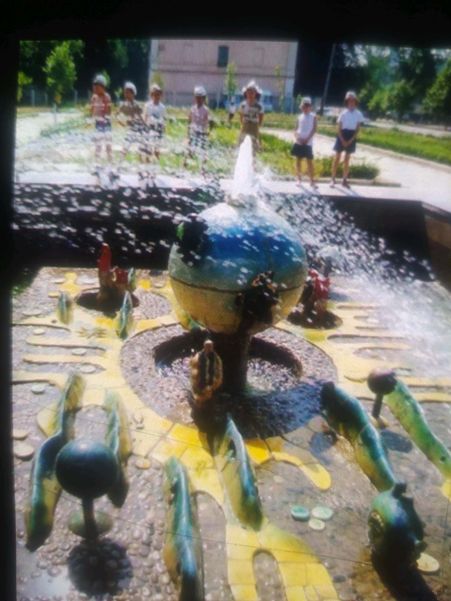 The image depicts a fountain with several children standing around it. The fountain has a globe-like structure at its center, with water spraying upwards. Around the base of the fountain, there are several colorful, abstract sculptures that appear to be part of the fountain's design. The ground surrounding the fountain is paved with stones and decorated with yellow painted lines. In the background, there are trees, a building, and a pathway. The children are wearing casual clothing, and some appear to be interacting with the fountain. The scene is set outdoors on a sunny day. This scene is located in the city of Birlik, Uzbekistan.