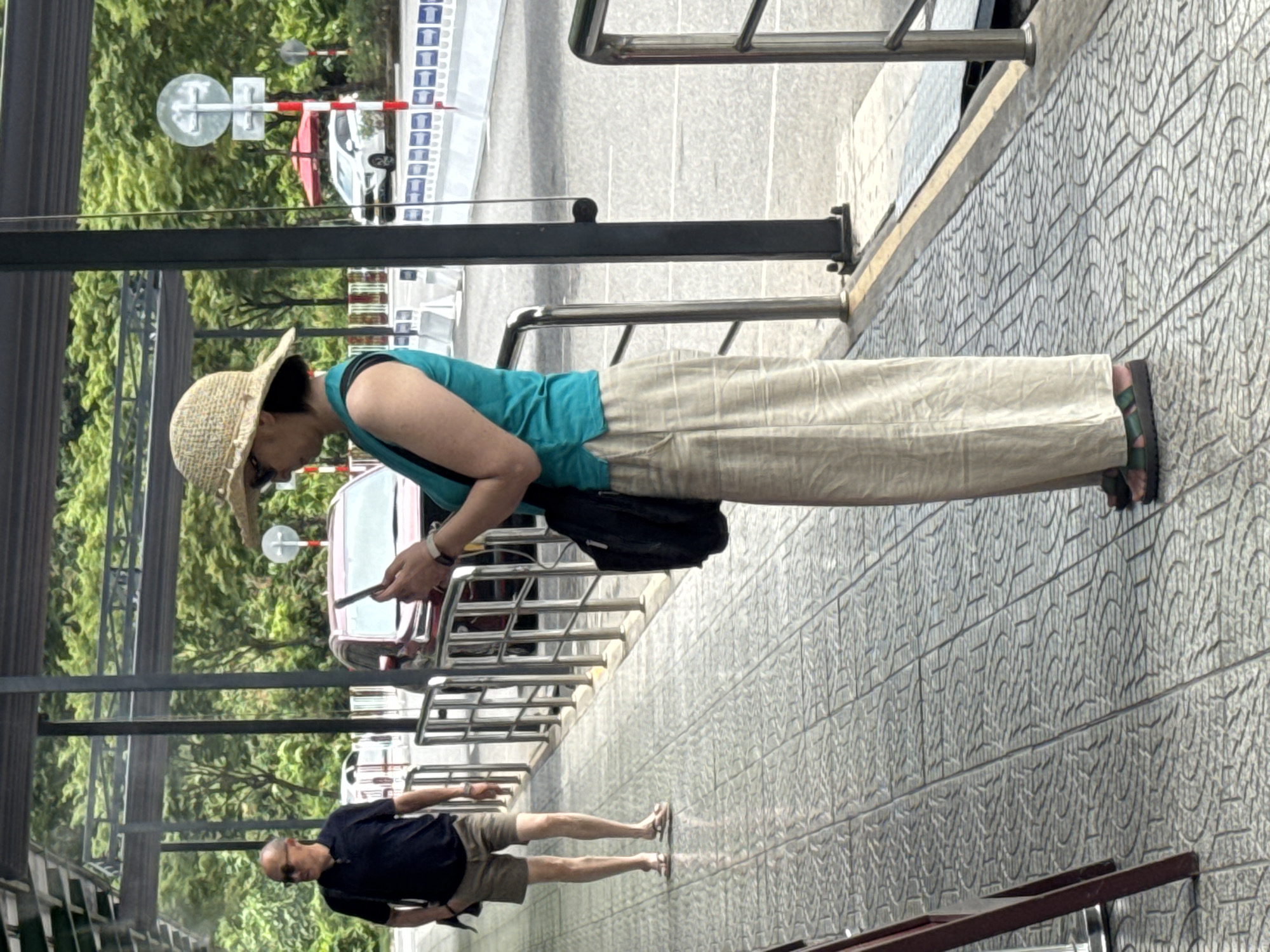 A medium, eye-level shot shows two people on what appears to be a pedestrian bridge. A woman in a straw hat, sunglasses, turquoise top, and khaki pants stands looking at her phone. She has a black bag slung over her shoulder. Below her, a man in a navy polo shirt and khaki shorts stands looking towards the left. They are standing on a textured grey surface with a metal railing next to them. Beyond the railing, there are trees and traffic signs. The location is assumed to be Đà Nẵng, Vietnam.