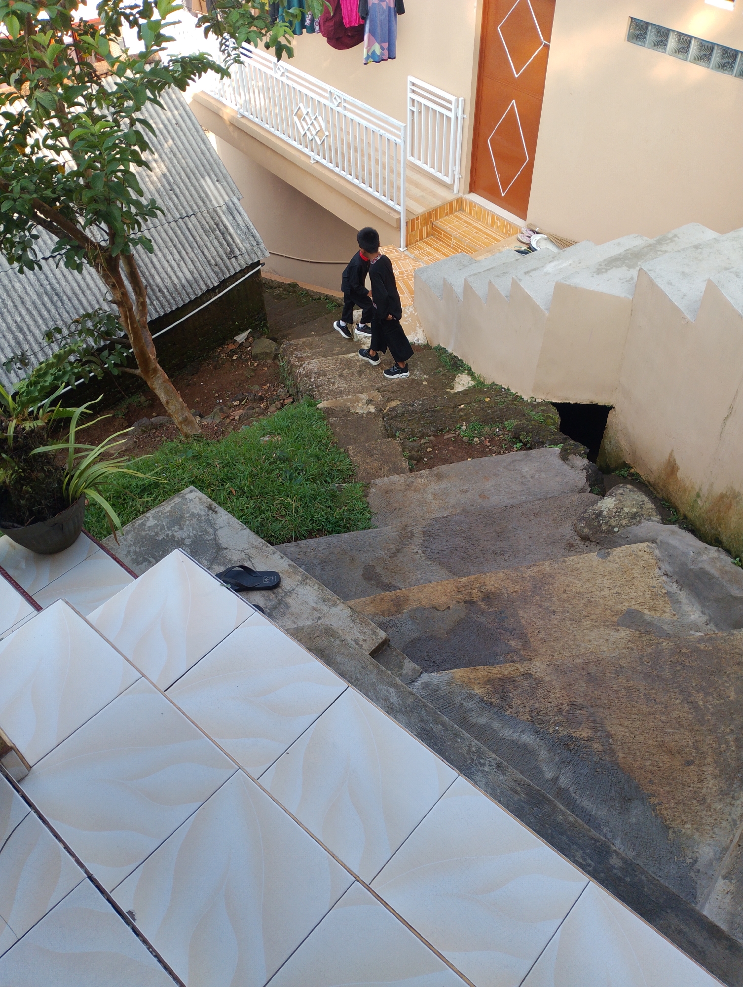Two young children, one wearing a black long-sleeved shirt and black pants, and the other a similar black outfit, are walking down a set of outdoor concrete stairs. They are positioned in the upper-middle section of the frame, walking away from the camera. To the left of the stairs, a small tree with green leaves and a thick trunk stands beside a corrugated metal roof. Further to the left, a potted plant with long green leaves sits near the base of the tree. A single black sandal is visible on one of the concrete steps in the foreground. The stairs are bordered by a tan-colored wall with a jagged top edge on the right. A dark opening, possibly a drain or small tunnel, is visible in the wall at the bottom of the stairs. The scene appears to be an outdoor residential area, possibly within the city of Sukamanah, Indonesia, characterized by its utilitarian construction and vegetation.