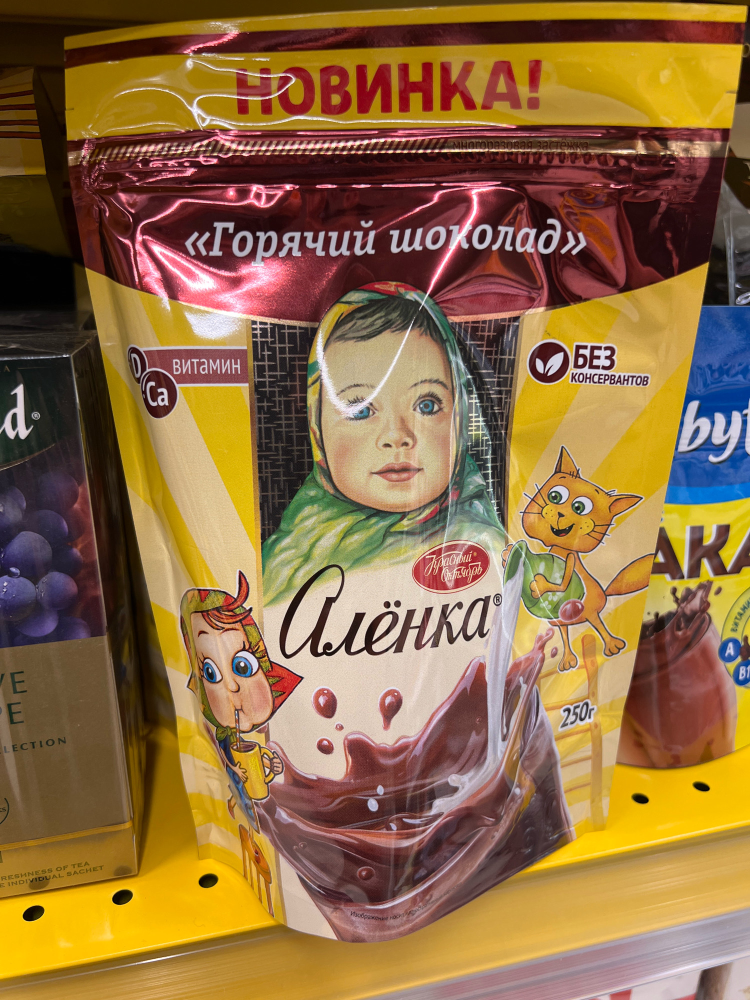 The image shows a close-up of a product package of "Alyonka" brand hot chocolate. The packaging is predominantly yellow with red and brown accents. A stylized illustration of a baby wearing a headscarf is featured prominently in the center, with a cartoon cat to its right pouring milk into a mug. Text on the package includes "НОВИНКА!" (New!), "«Горячий шоколад»" (Hot Chocolate), and "БЕЗ КОНСЕРВАНТОВ" (Without Preservatives). There are also labels indicating "ВИТАМИН D" (Vitamin D) and "Ca" (Calcium). The product weighs "250г" (250g). The packaging is slightly crinkled, suggesting it is a flexible pouch. The product is displayed on a yellow shelf, with other products partially visible to the left and right. The image appears to be taken in a retail environment, likely a grocery store shelf in Tenki, Russia.