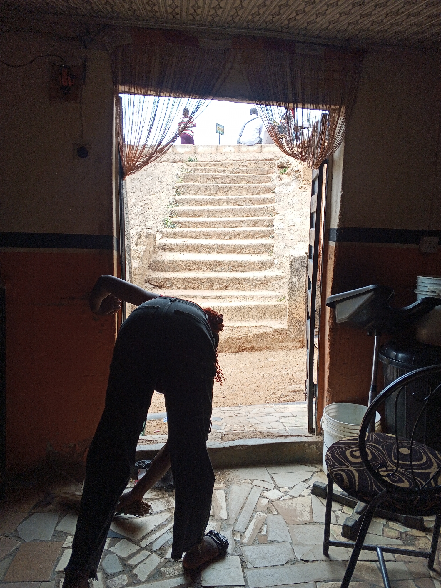 A person bends over, sweeping a mosaic tiled floor inside a building in Gwong, Nigeria. The doorway behind them leads to a set of exterior stairs. Two people are sitting at the top of the stairs. A chair and hair salon equipment are visible in the room. The doorway is draped with string curtains.