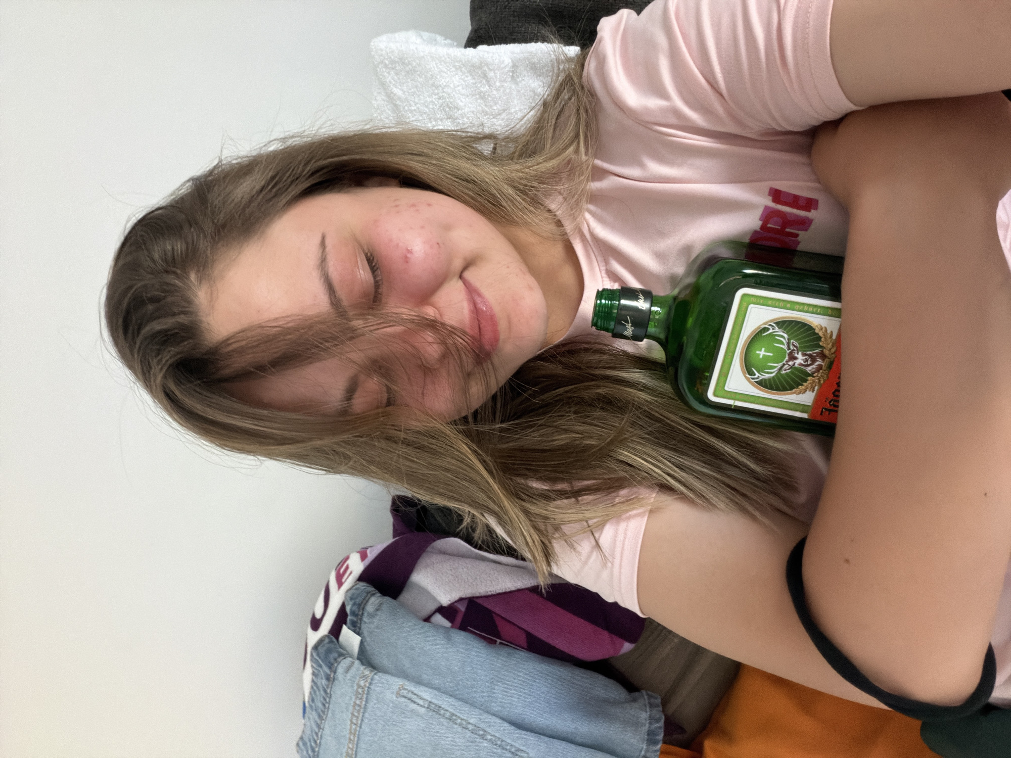 A young woman is reclined, possibly lying down, in an indoor setting. She is wearing a light pink t-shirt and has brown hair. She is holding a green glass bottle of Jägermeister. The background shows various clothing items, including a white towel and a blue denim garment. The photo does not provide enough information to determine a specific location in Arroyo de la Miel-Benalmádena Costa, Spain.