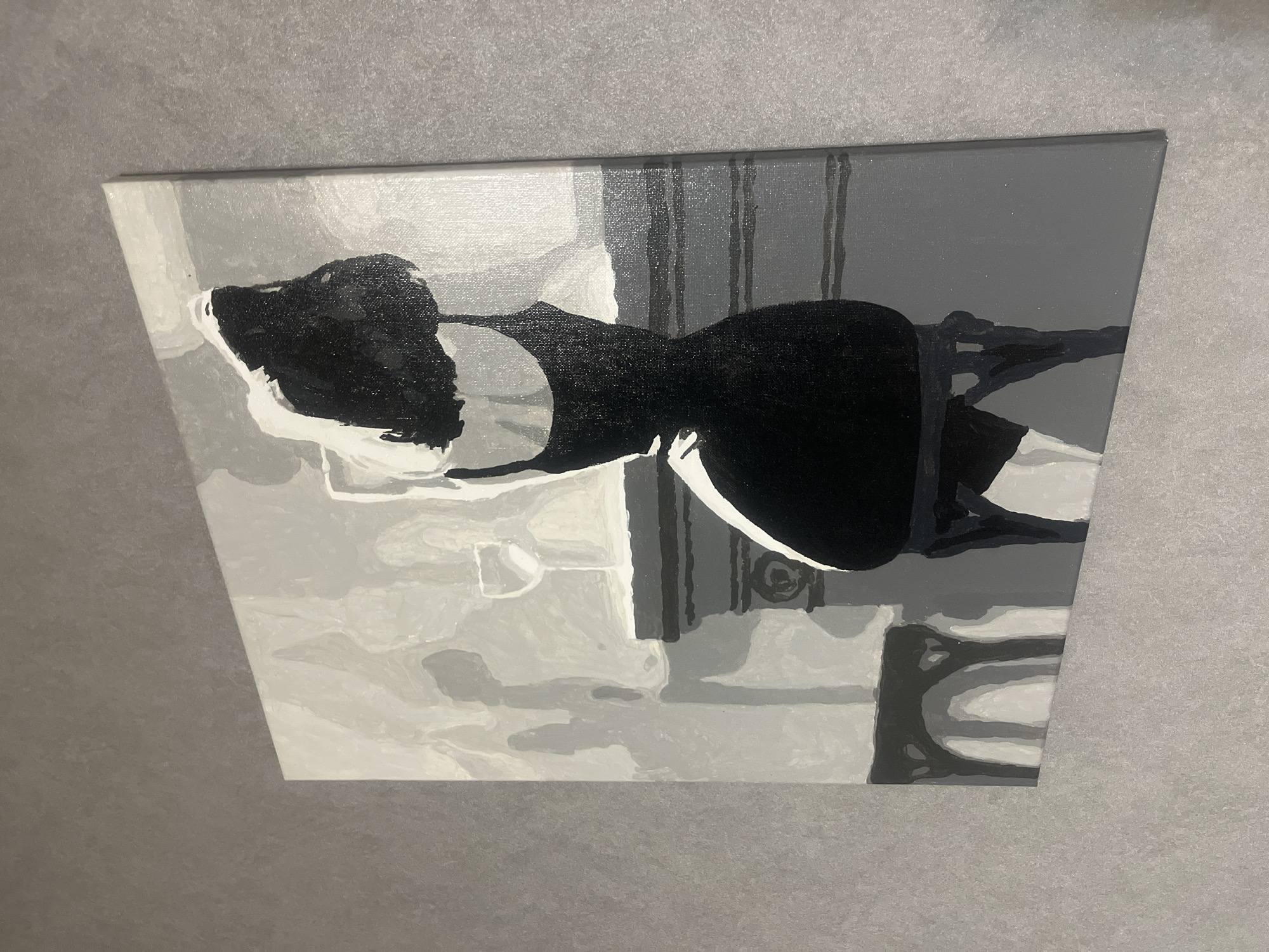 A monochrome painting, depicting the back of a woman in a black dress, is mounted on a textured gray wall. The painting is a stylized representation with broad strokes and a limited palette of black, white, and various shades of gray. The woman's form is central, her dark hair obscuring her face. She appears to be seated on a chair or stool, the legs of which are visible beneath her. In the background, there are suggestions of a mantelpiece with a wine glass and ornate decorative elements. The overall impression is one of subtle elegance and mystery. The specific location in the city of Rybnitsa, Moldova, is not determinable from the visual information provided in the image.