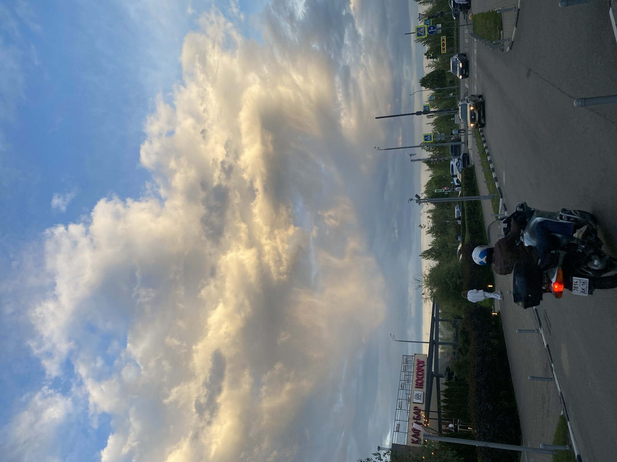 The image depicts an urban street scene in Kazan, Russia, during what appears to be late afternoon or early evening. The sky dominates the upper portion, featuring a mix of clear blue patches and large, textured clouds. Many of these clouds are dramatically illuminated with warm, golden light, indicating a low sun, while others appear in various shades of grey.
A paved road runs along the right side of the frame, with several vehicles present. In the foreground, a motorcycle with a person wearing a helmet is prominently visible, its tail light illuminated. The motorcycle's Russian license plate, "7814 AK 116", is clearly readable. Further down the road, multiple cars are seen, with at least one car's headlights turned on.
To the left of the road, a sidewalk or paved path is visible, bordered by a strip of grass and shrubs. A person dressed in white clothing is walking on this path, facing away from the camera. Further left, a building is partially visible, adorned with a large sign featuring Russian text: "КАФЕ-БАР" (Cafe-Bar) and "НОСОРОГ" (Rhinoceros). Numerous streetlights and yellow pedestrian crossing signs are situated along the roadside. The scene conveys a sense of a routine urban evening, enhanced by the vibrant and dynamic cloud formations.