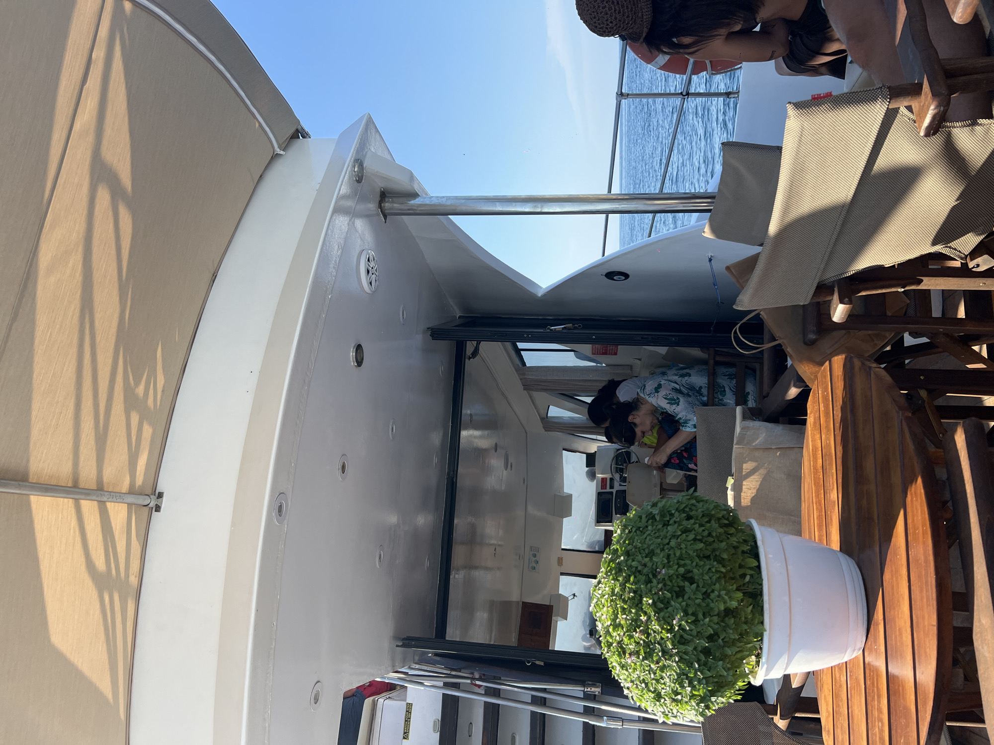 The image depicts a scene on the deck of a yacht, likely docked or cruising in the Aegean Sea, near the island of Skopelos, Greece. In the foreground, a round wooden table is set with a white pot containing a lush green plant. Behind the table, several empty chairs with beige fabric are arranged.
The yacht's structure is prominent, with a light-colored awning casting shadows over the deck. A metal railing runs along the edge of the deck, with the blue sea visible beyond. In the mid-ground, an open doorway leads into the yacht's interior, where several people are seated. One person is at the helm of the boat, with a steering wheel and various controls visible.
To the right, a woman wearing a hat is partially visible, facing away from the camera. A life preserver is hanging on the railing. The overall ambiance suggests a leisure activity on a sunny day.