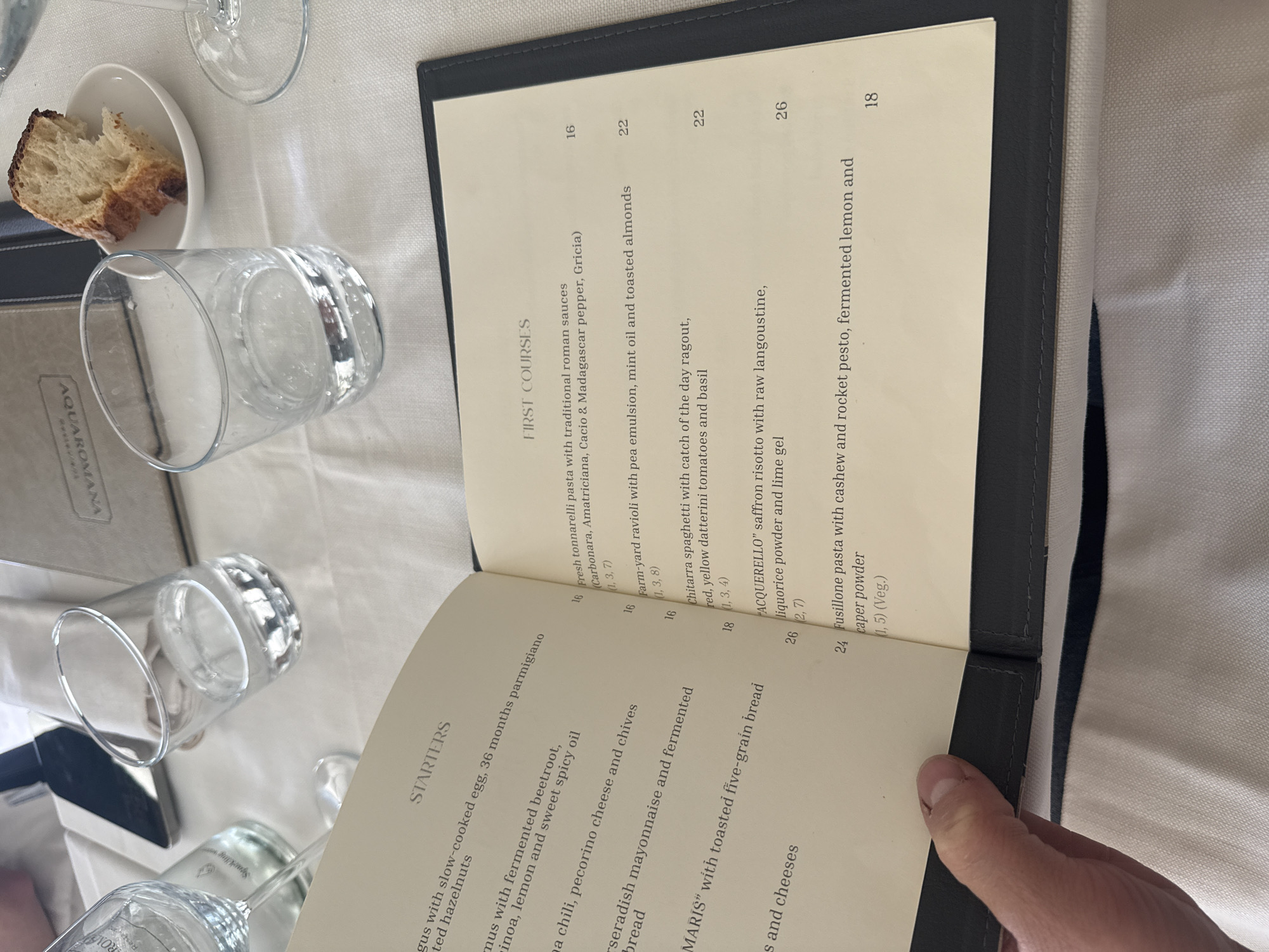The image shows a menu at Aquarolavagna restaurant in Bracciano, Italy. The menu is open to the "Starters" and "First Courses" pages, listing various dishes and prices. A hand is holding the edge of the menu. In the background, there are several glasses, a piece of bread in a small dish, and a table setting. The table has a white tablecloth.