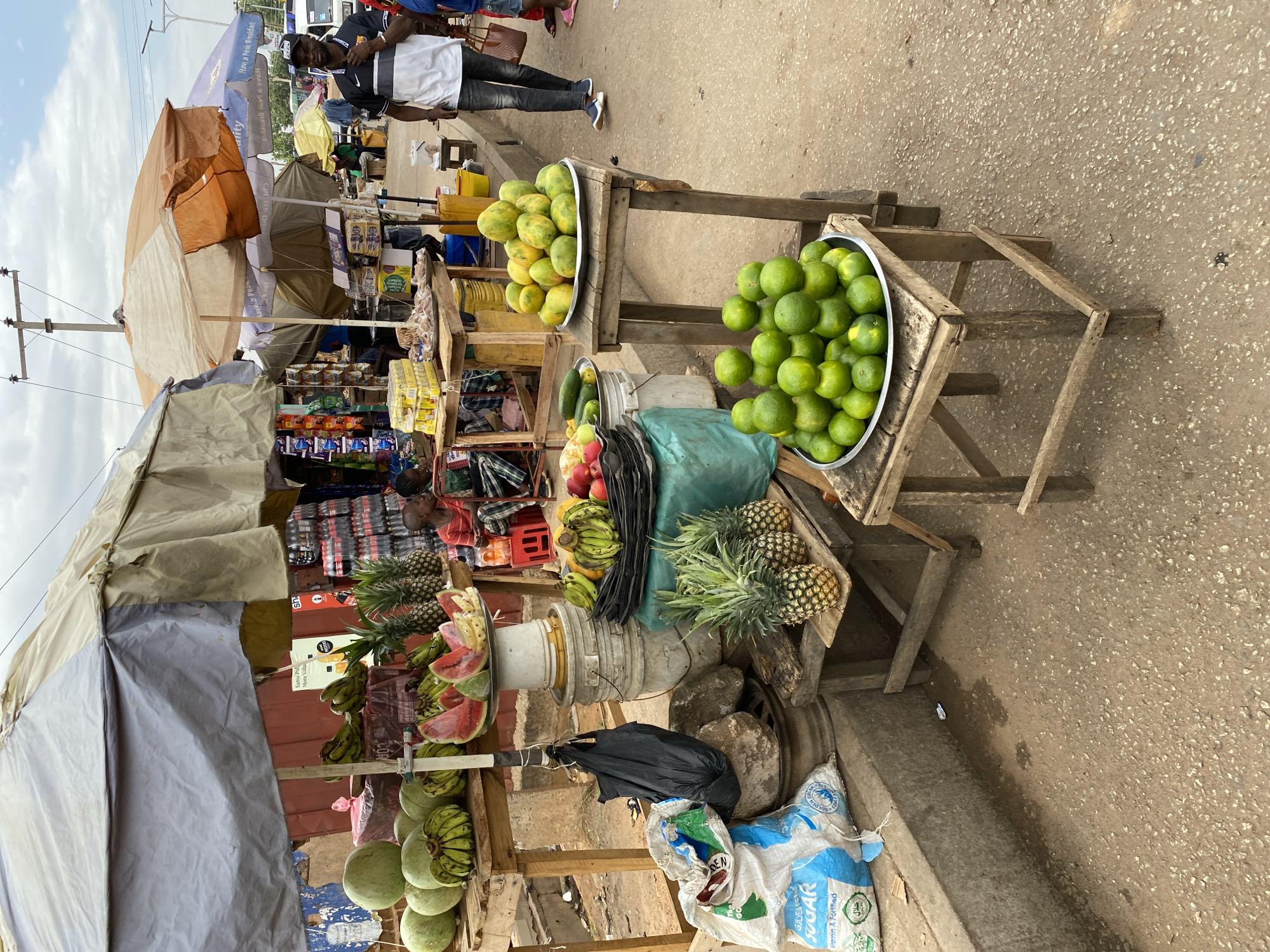 The image captures a roadside fruit and goods market in Bukuru, Nigeria. Vendors display produce such as limes, mangoes, pineapples, bananas, and watermelons on wooden stands. Packaged goods, including stacks of bottled drinks, are visible on shelves beneath the canopies, alongside bags of sugar. People are seen walking or standing nearby. The market appears to be set up along a paved street, with additional canopy umbrellas visible in the background.