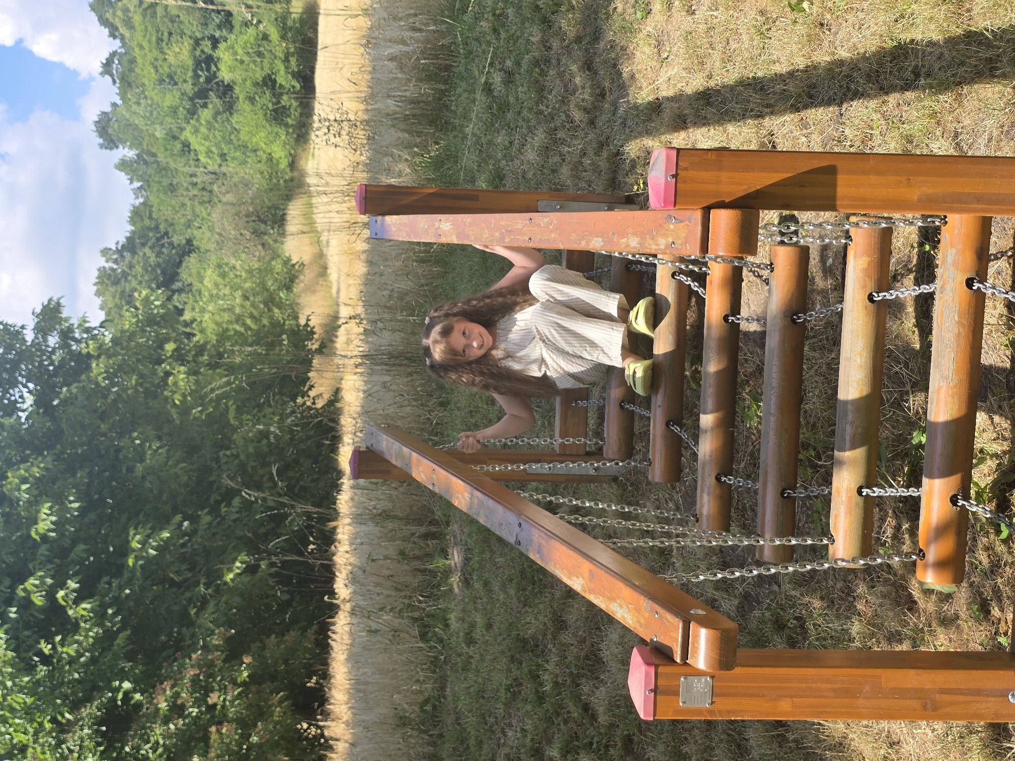 In Pustiměř, Czechia, a young girl with long, wavy brown hair styled in two pigtails sits on a wooden and chain playground obstacle. She is wearing a light-colored, striped jumpsuit and yellow slip-on shoes. The obstacle consists of wooden beams connected by metal chains, forming a ramp-like structure. The girl is smiling and looking directly at the camera. She is positioned on the upper half of the obstacle, with her hands resting on the wooden frame. The background shows a grassy area with dry grass and a field of wheat or similar grain, bordered by a dense line of trees. The sky is blue with scattered white clouds. The lighting suggests it is a sunny day.