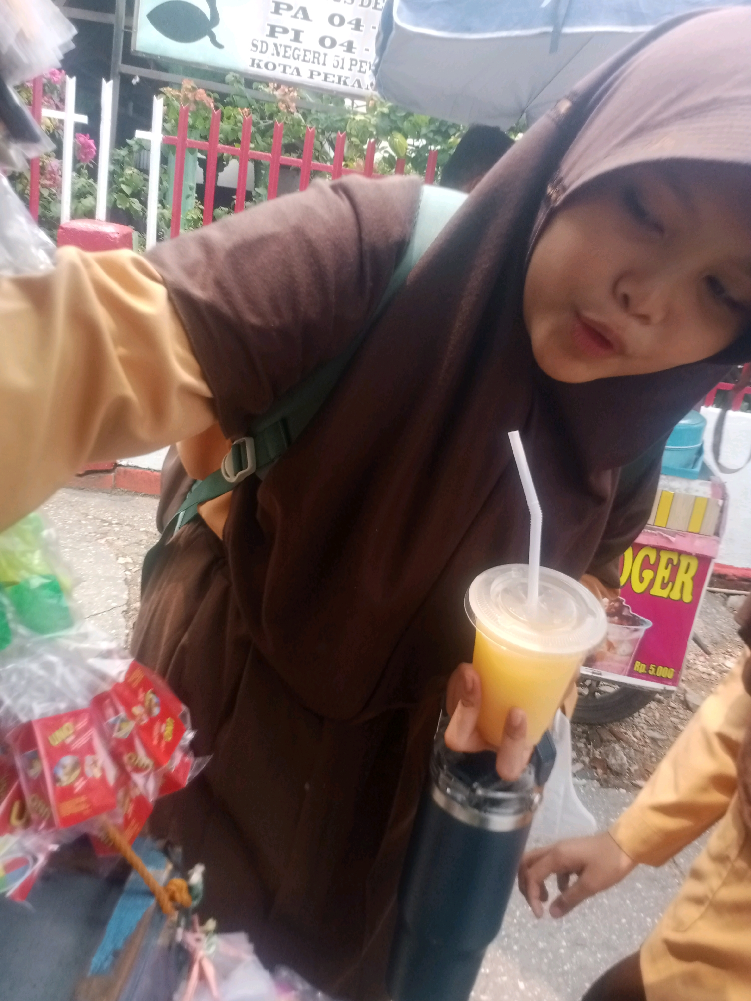 In a street scene in Pekanbaru, Indonesia, a young girl wearing a brown hijab and a tan long-sleeved shirt is holding a cup of orange liquid with a straw. To her left, several red packaged snacks are visible, some partially obscured by plastic. A dark blue tumbler is held in her other hand. In the background, a food cart with the word "OGER" in yellow lettering on a pink sign is present, along with a sign indicating "Rp. 5.000". A metal fence with white and red posts borders the area, and greenery with pink flowers can be seen behind it. The ground is paved with gravel and asphalt.