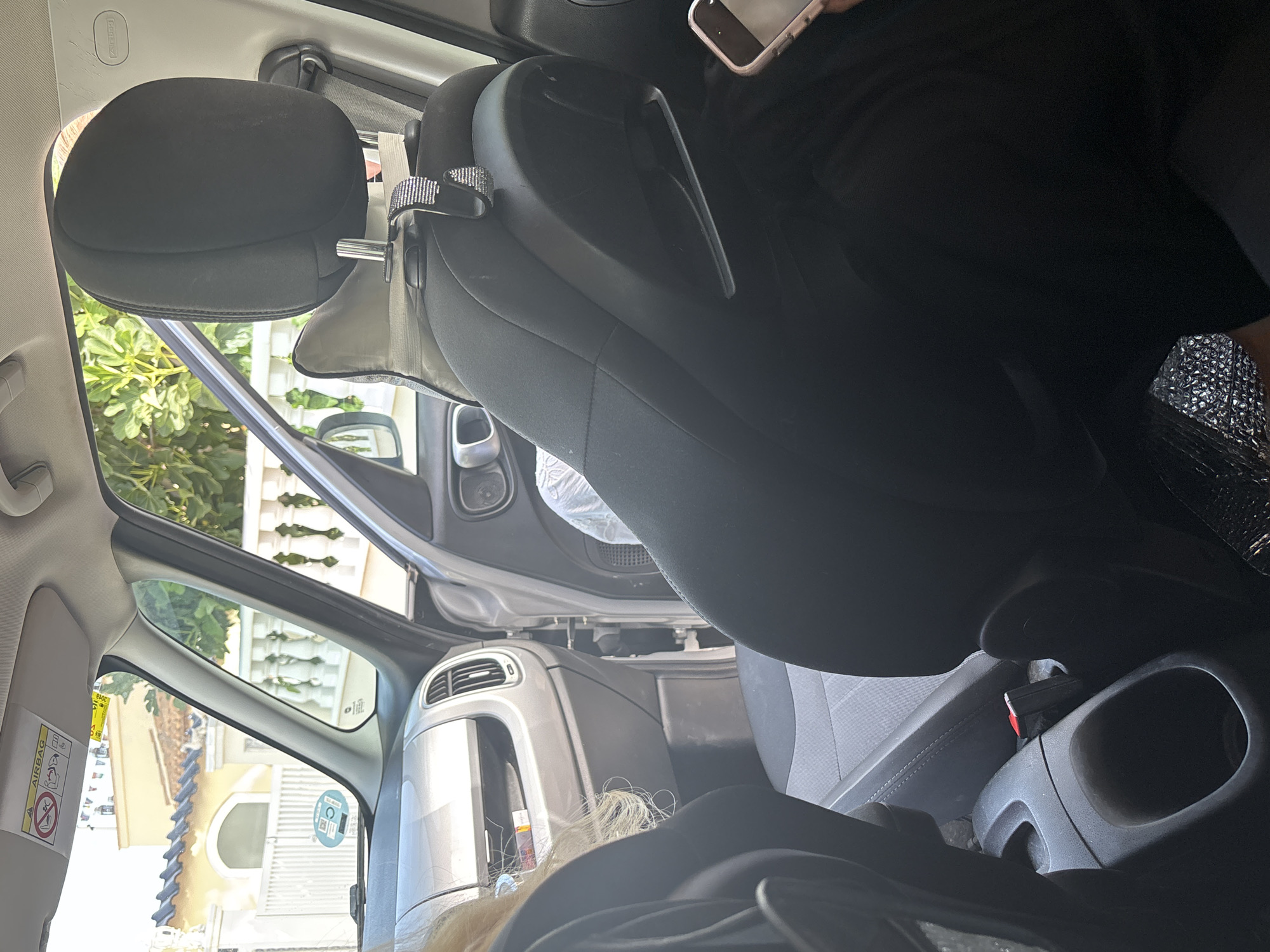 The image shows the interior of a car, viewed from the back seat, looking forward. The car's interior is predominantly black and grey. In the foreground, the backs of the rear seats are visible, along with a cup holder between them. A person's blonde hair is visible on the left side of the frame, and a phone is held by a person on the right. The front passenger seat is visible, with a headrest and part of the seat itself. Attached to the headrest is a sparkly hook. The dashboard is visible in the upper left of the image, with a sun visor above it. Outside the car, a building with yellow walls and a tiled roof can be seen, along with some greenery and a white balustrade. The location is unknown.