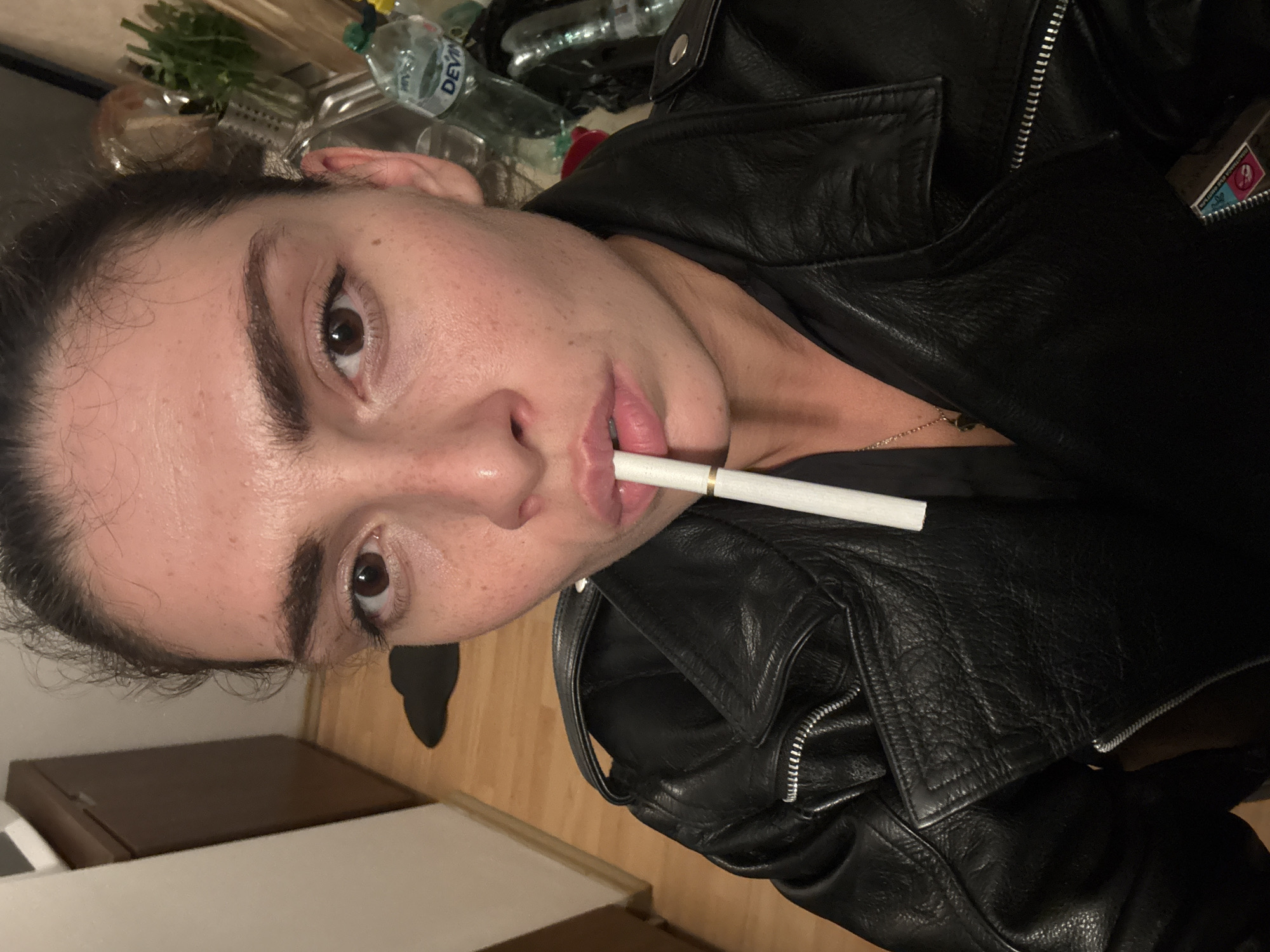 A close-up, eye-level portrait shows a person, likely female, with dark hair and prominent dark eyebrows, looking directly at the camera. The image is rotated 90 degrees clockwise. She has a fair complexion with some visible freckles or blemishes. A white cigarette or vape pen is held between her slightly parted lips.
The subject is wearing a black leather jacket with silver zippers and snaps, suggesting a biker or stylish design. A delicate gold chain necklace is visible around her neck.
The background suggests an indoor setting, possibly a kitchen or living area, with artificial lighting. Behind her, a plastic water bottle with the brand "DEVIN" clearly visible is placed on a surface, alongside some greenery in a container. Another blurred plastic bottle is also present. Below the person, a wooden surface, possibly flooring or furniture, is visible, along with a dark brown rectangular object. Tucked into a crevice or pocket of the leather jacket, a small rectangular object with the number "9" prominently displayed is partially visible. The "DEVIN" brand on the water bottle is a strong indicator of the location being Sofia, Bulgaria, as provided in the context. The overall mood is direct and somewhat serious.