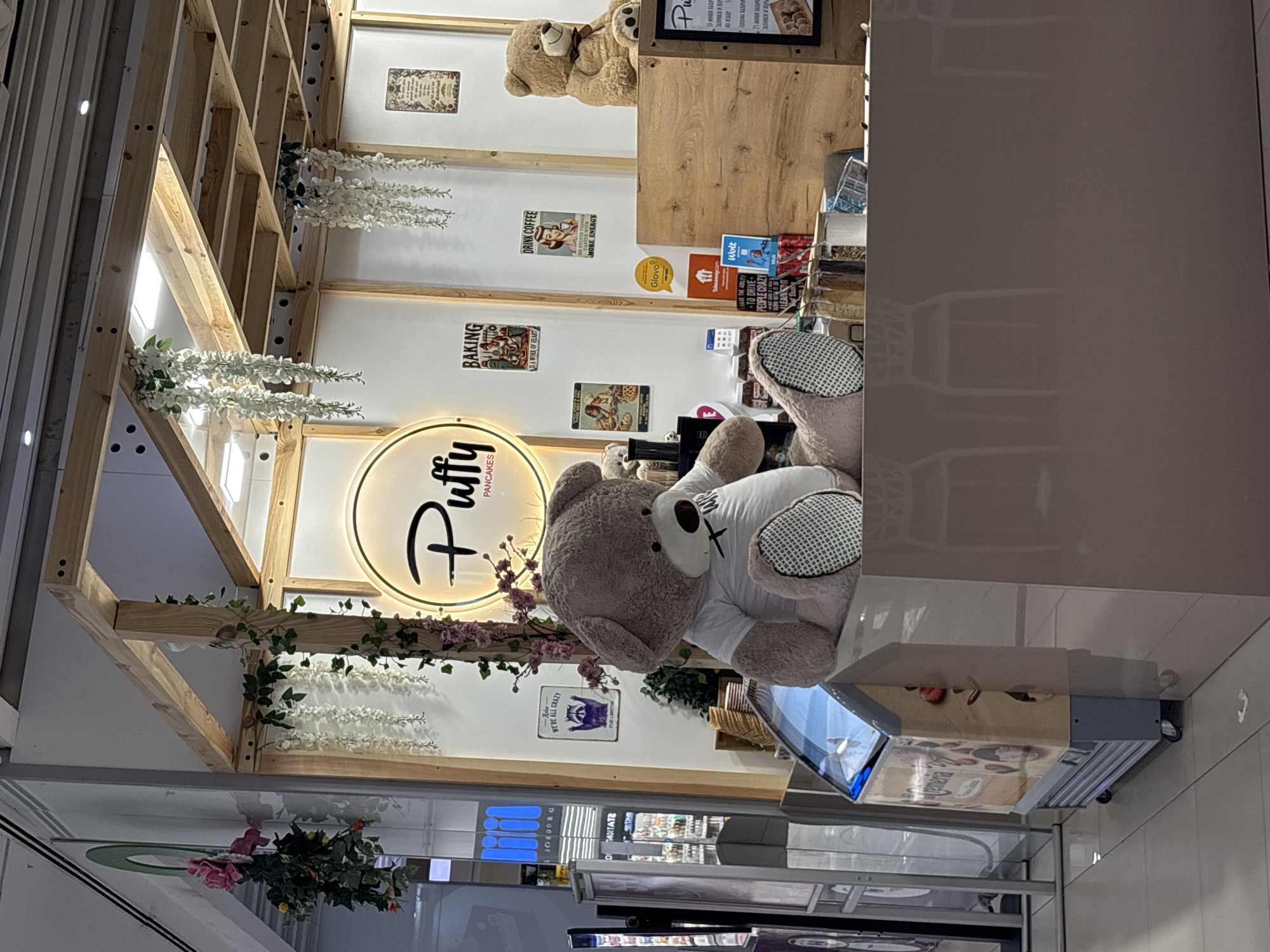 The image shows a "Puffy Pancakes" stall, likely located inside a shopping mall. The stall is decorated with wooden beams and artificial flowers. A large teddy bear wearing a white shirt sits in front of the stall. The stall features a lighted sign with the brand name "Puffy Pancakes," along with framed images on the back wall depicting pancakes, baking scenes, and coffee advertisements. Other details include a counter area with order placement screens, delivery service logos, and signage with prices. The location is likely Plovdiv, Bulgaria, based on the text "ЗАЛИВКИ И ПОР" on the signage.