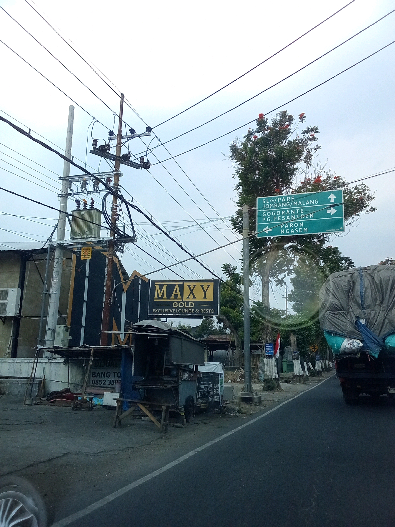 The image captures a roadside scene in Ngasem, Indonesia, with a focus on electrical infrastructure and directional signage. A prominent utility pole with a transformer and numerous wires dominates the left side of the frame. Adjacent to it, a building with the signage "MAXY GOLD EXCLUSIVE LOUNGE & RESTO" is visible. A street vendor stall is situated in front of the building.
To the right of the utility pole, a green directional signpost displays several locations: "SLG/PARE JOMBANG/MALANG" with an upward arrow, "GOGORANTE PG.PESANTREN" with a rightward arrow, and "PARON NGASEM" with a leftward arrow. These signs indicate directions to various towns and areas.
The road itself is paved and appears to be a two-lane street. A large truck, partially covered with a tarp, is driving on the right side of the road. The environment is characterized by trees lining the street, some with red flowers, and a cloudy sky. The ground is paved in some areas and appears to be dirt or gravel in others, particularly around the vendor stall. The overall scene depicts a typical Indonesian road with functional infrastructure and commercial activity.