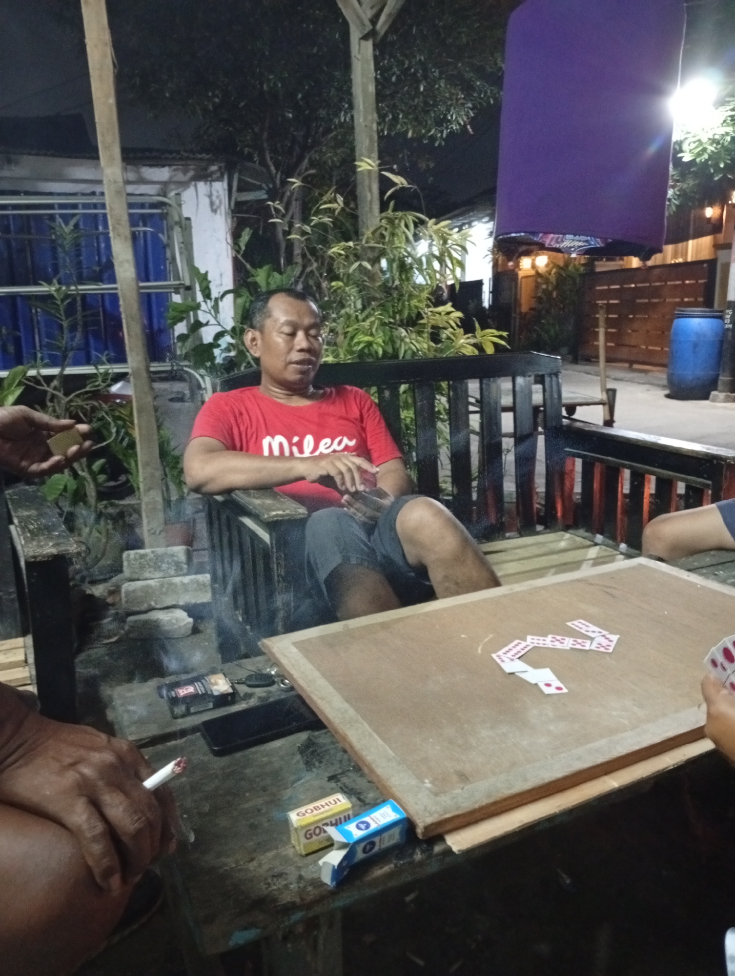 The image depicts an outdoor setting at night, possibly in a relaxed social environment in Caringin, Indonesia. The foreground shows a wooden table with a game in progress, indicated by several dominoes laid out. Next to the dominoes are two small boxes, one labeled "GOBHUI GOSHUI". A hand is visible on the right side of the frame, holding a set of dominoes. In the mid-ground, a man wearing a red t-shirt with the word "Milea" printed on it is seated on a wooden bench. He appears to be looking down and is smoking a cigarette. Another person's hand holding a cigarette is visible in the lower left corner of the frame, with ash falling onto the table. Behind the man, a wooden bench with a dark frame and lighter wooden slats is situated. The environment is dimly lit by artificial light, with some vegetation and a building visible in the background. A blue plastic barrel and a wooden fence are also present in the background. The overall atmosphere suggests a casual gathering, possibly in a yard or an open-air establishment.