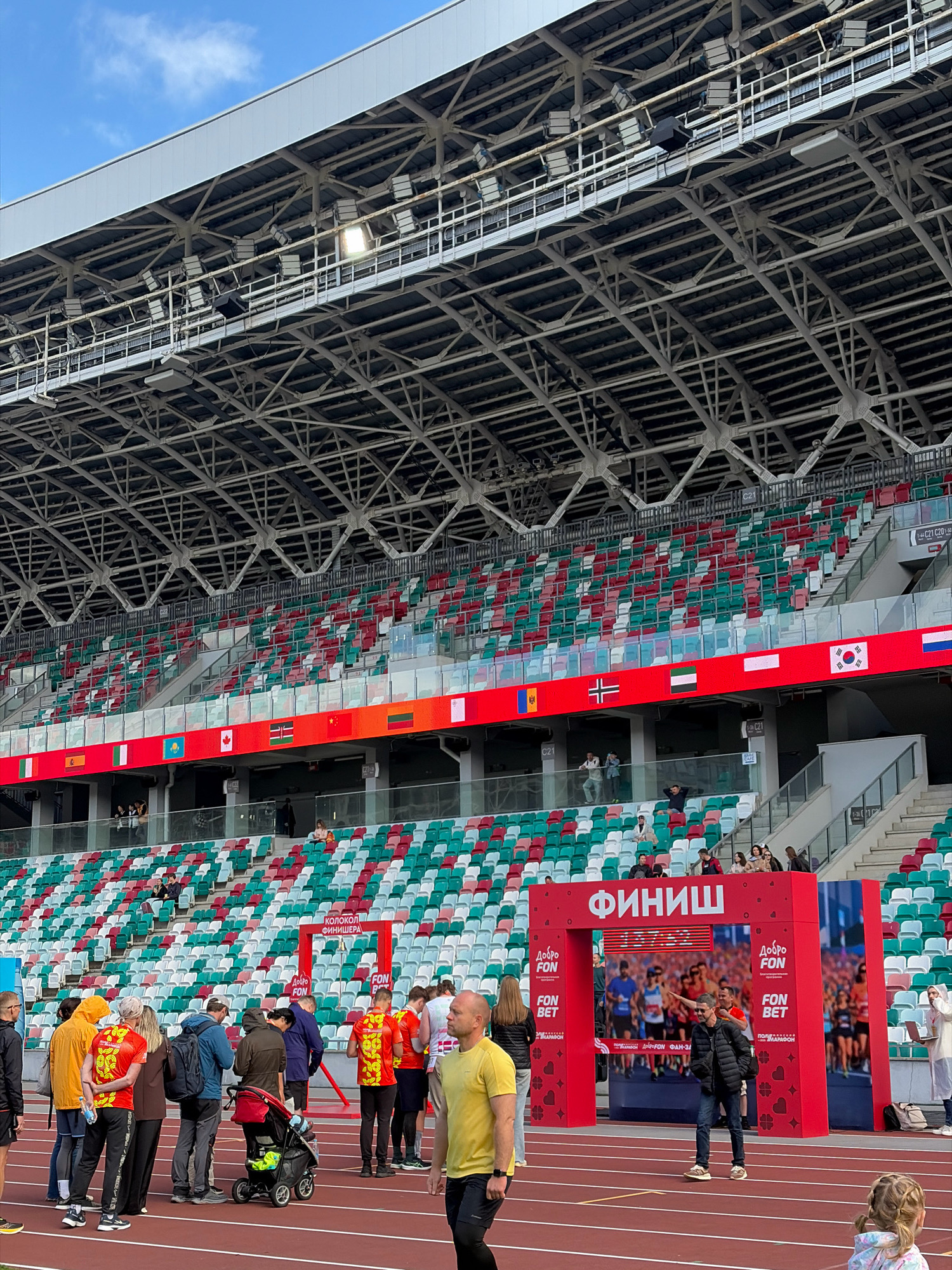 This is a wide shot of an outdoor athletics stadium, likely in Minsk, Belarus, during an event. The stadium seating is a mix of red, green, and white seats, arranged in sections. A prominent red archway with "ФИНИШ" (Finish) written in large white Cyrillic letters stands on the track. The archway is adorned with sponsor logos, including "ДОБРО FON" and "FON BET". Behind the archway, a large screen displays an image of runners. Several people are gathered on the track in front of the archway, some wearing brightly colored running attire. Some are looking at the screen or talking to each other, while others are pushing a stroller. The track itself is a standard red athletic surface. In the background, above the seating, is a large, complex metal structure supporting stadium lights and the stadium roof. The sky is cloudy.