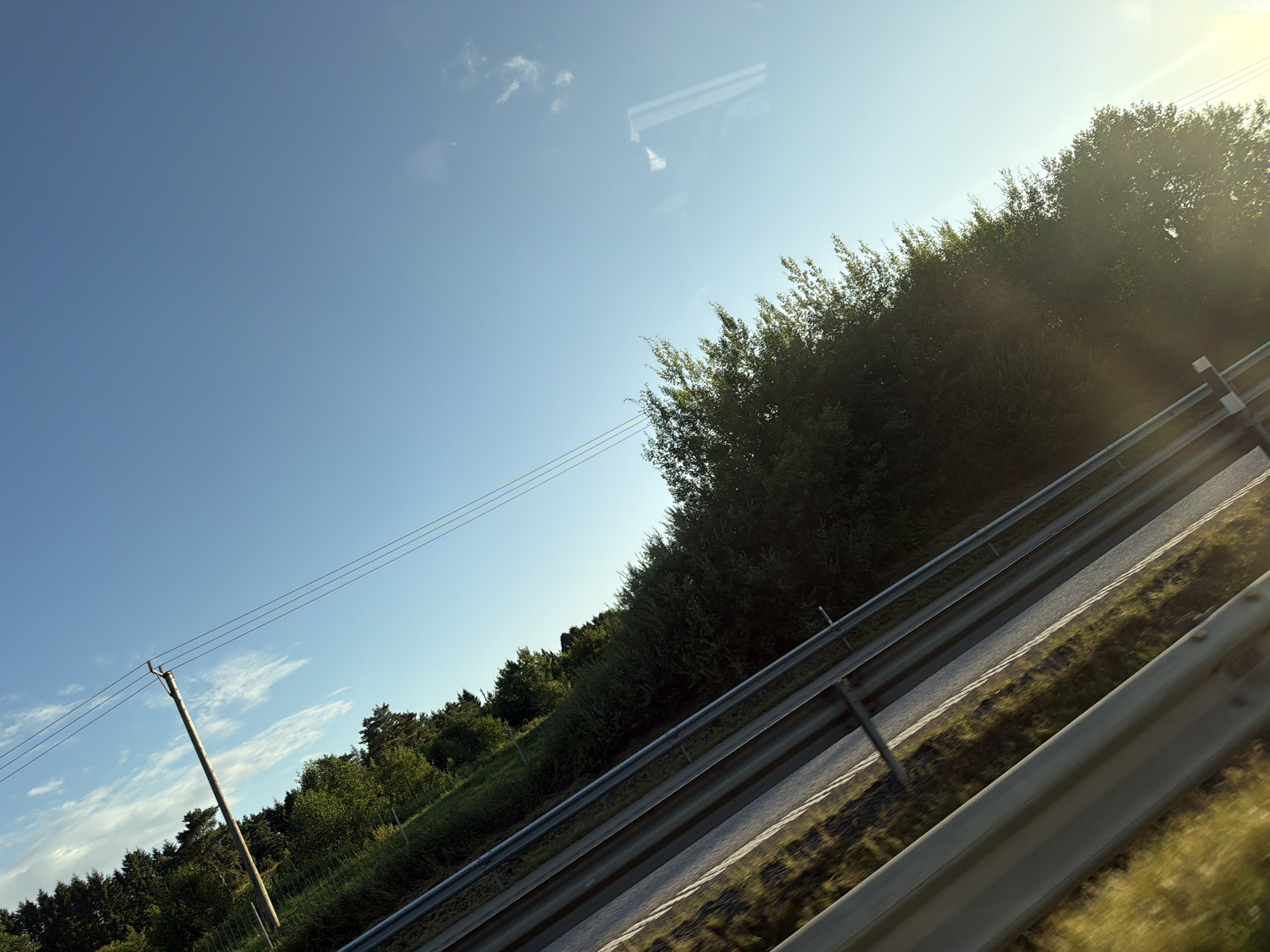 The image shows a view from a car traveling on a road in Kareby, Sweden. The road is bordered by a metal guardrail on the left, with a grassy shoulder and dense trees on the right. A utility pole with three wires is visible on the left side of the road, against a clear blue sky with a few wispy clouds. The sun is low in the sky, casting a warm light and some lens flare. The perspective suggests the photo was taken from inside a moving vehicle, possibly looking out the window.