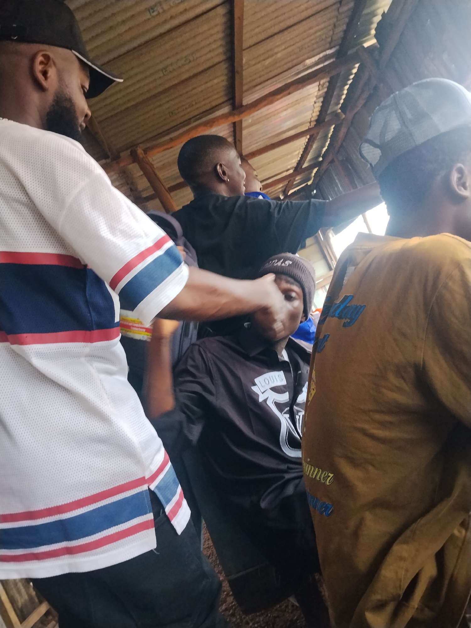 In Durumi, Nigeria, under a corrugated metal roof supported by wooden beams, four individuals are gathered. The person closest to the viewer is wearing a white mesh jersey with red and blue horizontal stripes on the sleeves and hem. To their left, a man with a beard wears a black baseball cap. In the center, a person wearing a black beanie and a black long-sleeved shirt with a white logo is partially obscured, with another individual in a yellow-brown jacket standing to the right, partially blocking the view of the center subject. The overall scene depicts an informal gathering in an open-air structure.