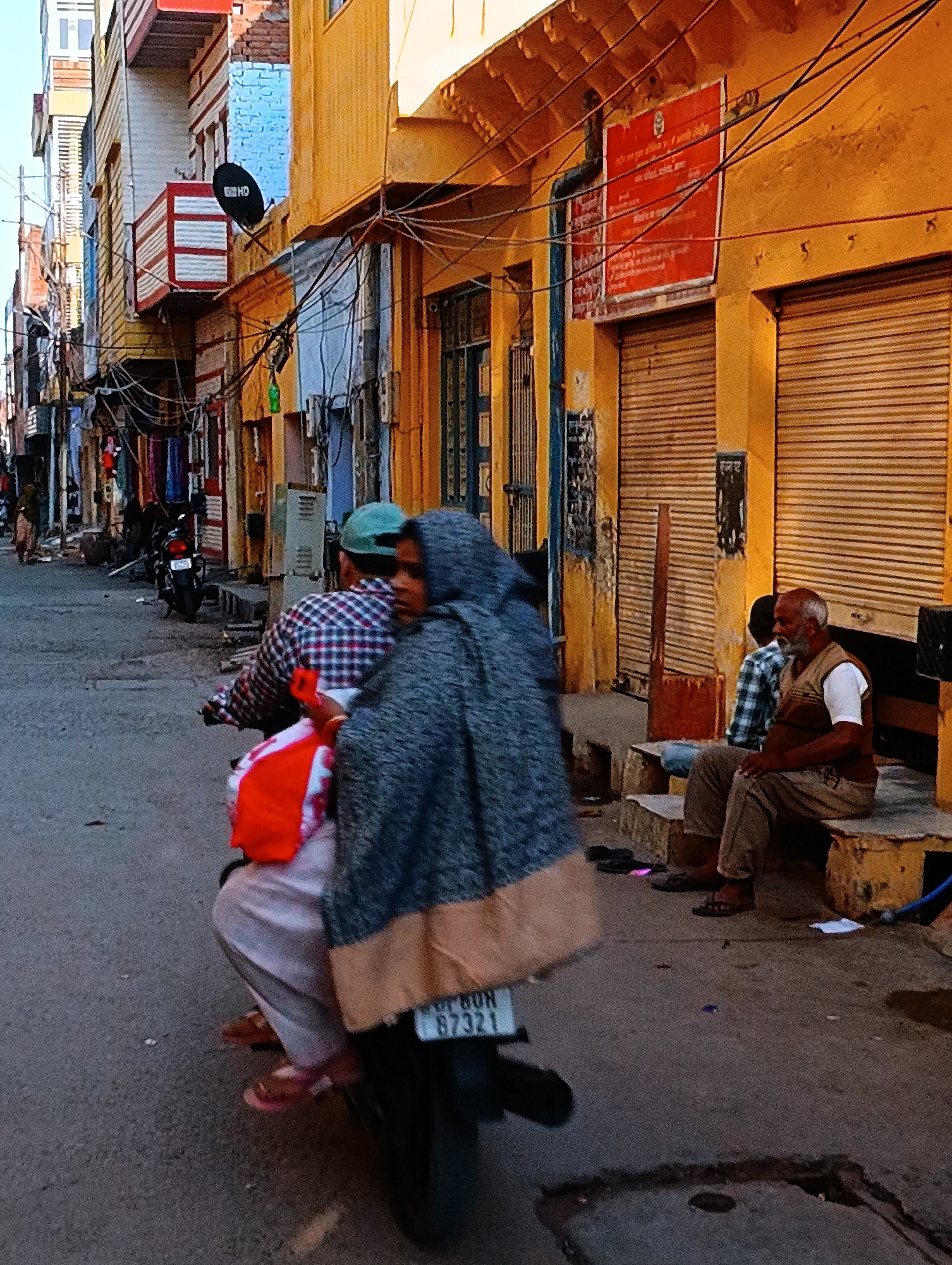 The image depicts a street scene in an unidentified city in India. A man wearing a plaid shirt and green cap drives a motorcycle with a passenger covered in a grey and tan shawl. The motorcycle license plate reads "RJ 16 DN 87321." On the right side of the street, an elderly man with a white beard sits on a concrete step next to a young boy wearing a plaid shirt. Behind them, two storefronts have closed metal shutters. A red sign is mounted above the storefronts. Overhead, a dense network of electrical wires crisscrosses the street. The buildings are painted in shades of yellow, blue, and white, with some featuring balconies and satellite dishes. Other pedestrians and motorcycles are visible in the distance along the street.