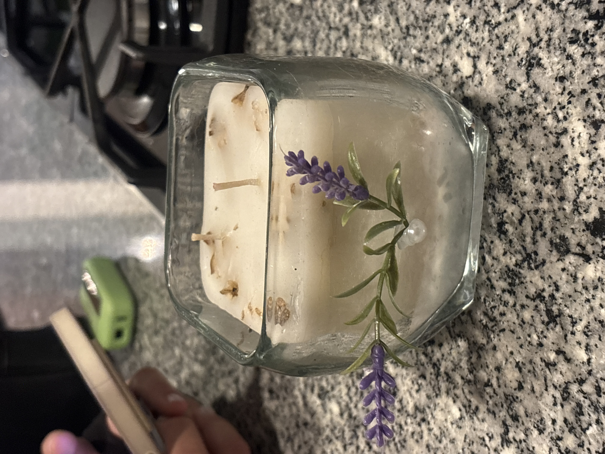 The image, taken from an overhead perspective and rotated, captures a clear glass container holding two white, square-shaped candles on a speckled countertop. The candles are embedded with what appear to be dried botanical pieces, possibly fragments of herbs or flowers. Attached to the side of the container, positioned between the two candles, is a decorative sprig of artificial lavender and small green leaves, accented by a single white pearl bead. Below this artificial sprig, a larger artificial lavender bloom hangs downwards.
The countertop, visible around the container and extending into the background, is a light gray or white granite/quartz with dark black and gray specks. In the blurred background to the left, a dark, angular object is partially visible, likely part of a kitchen appliance such as a stove or microwave oven. A small, out-of-focus light green stapler is also visible in the mid-left background. In the lower left foreground, a person's hands are visible, holding a light-colored smartphone or similar device. The scene appears to be an indoor domestic setting, possibly a kitchen. There is no visible text in the image.