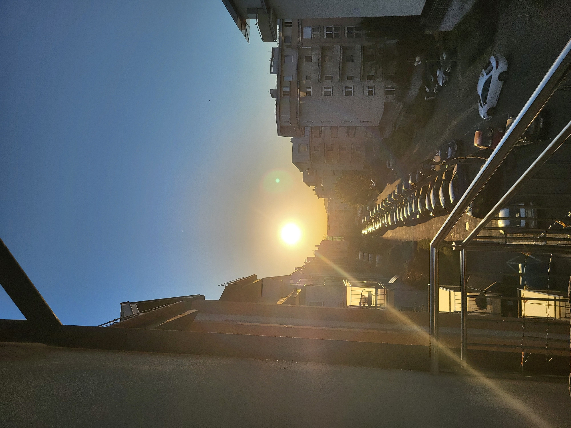 The image shows a street in an urban setting at sunrise or sunset. Tall buildings are on both sides of the street, and cars are parked along the right side. The sun is shining brightly in the distance, causing a lens flare. A metal railing is visible in the foreground. The location is Unknown, Unknown.
