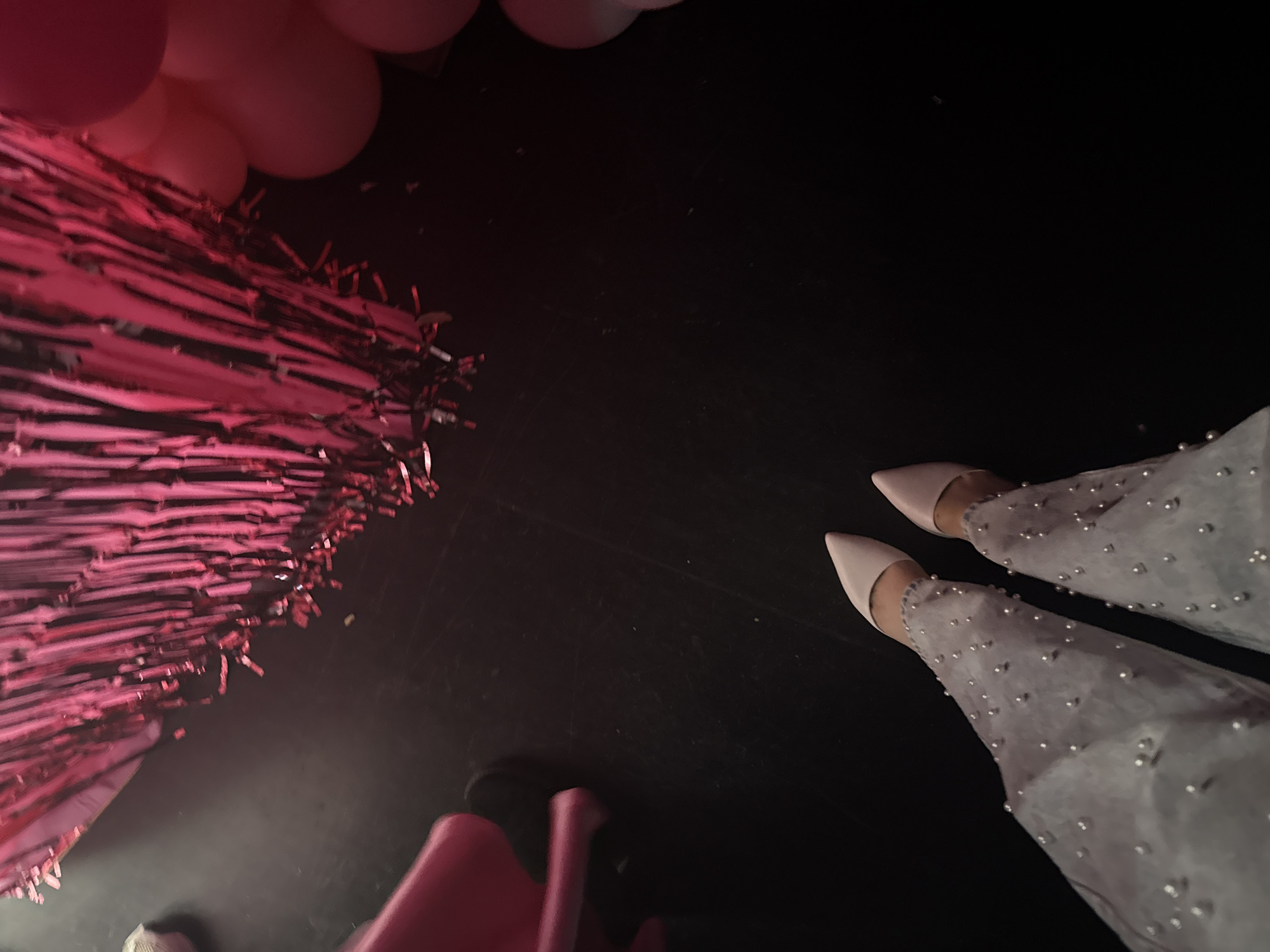 A high-angle shot shows a scene with party decorations and a person's legs. In the upper left corner are pink balloons and shiny pink fringe decorations. The background is a dark, likely black, surface, possibly a floor. In the lower right, a person's legs are visible, clad in light-colored, sheer pants adorned with silver beads or embellishments. The person is wearing pointy-toed, light pink or nude-colored high heels. In the lower left is part of a pink child's chair.
There is no information in the image that would indicate this took place in Plovdiv, Bulgaria.
