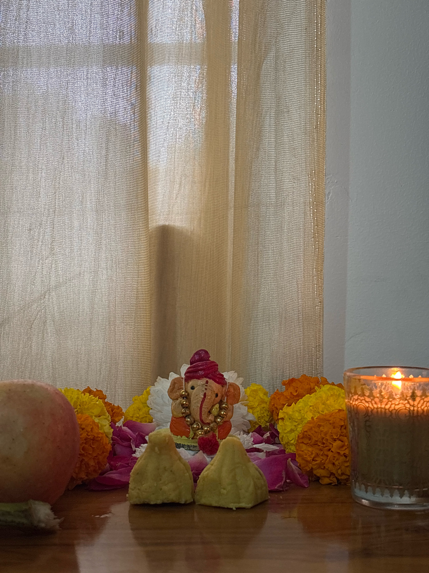 An outdoor, eye-level, medium shot captures a still life arrangement on a wooden surface in front of a sheer, beige curtain. The scene includes a small, ornamental figure of Lord Ganesha, adorned in a red turban and gold jewelry, positioned centrally. Flanking Ganesha are two cone-shaped modaks, a reddish-pink apple to the left, and a cluster of marigold flowers in shades of orange and yellow. Rose petals are scattered amongst the flowers. To the right, a lit candle in a glass holder emits a warm glow. The lighting suggests late afternoon or early evening. No people are present in the image, and there is no indication of the city of Bhatta, India.