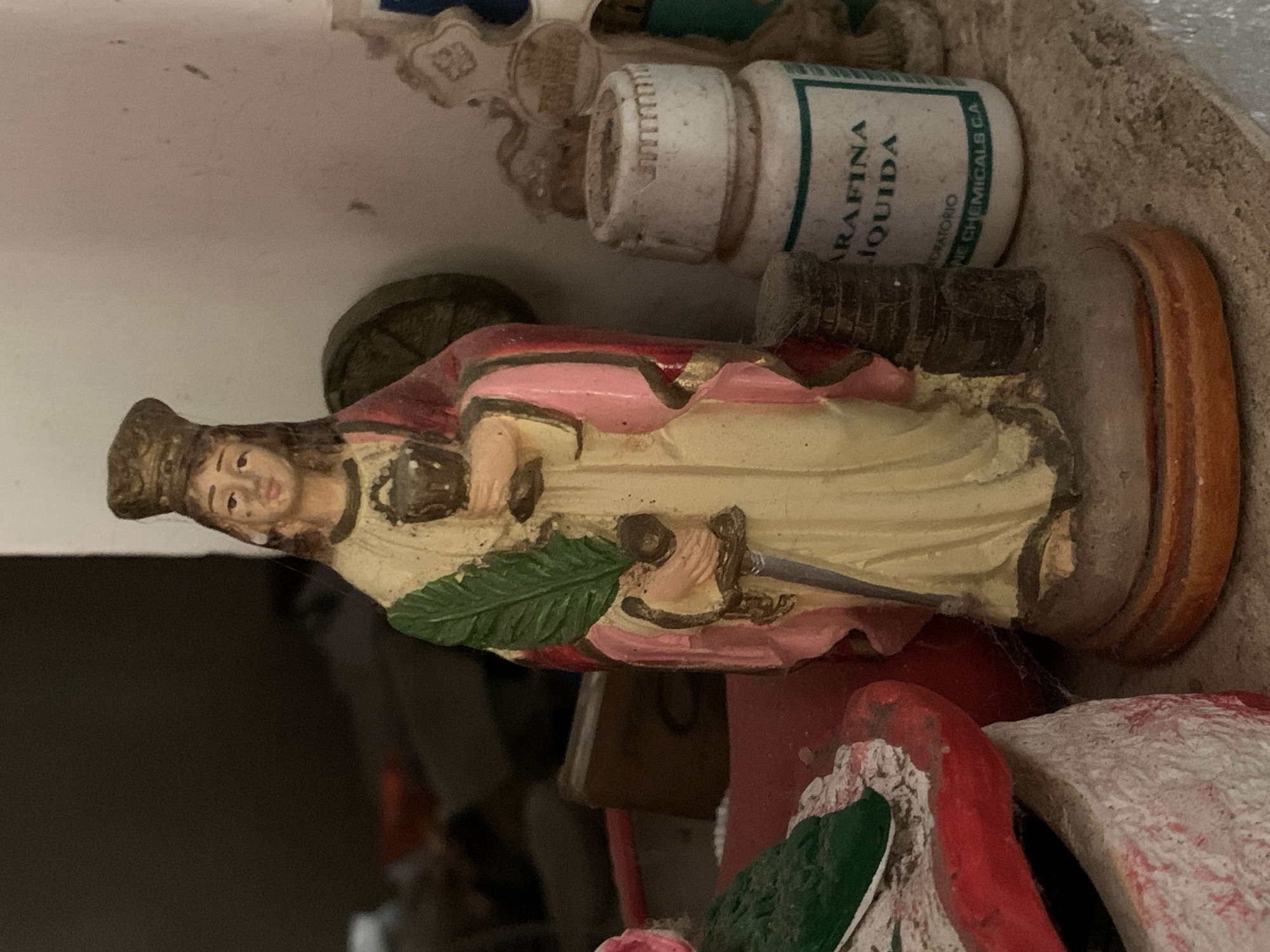 The image shows a close-up of a religious figurine, possibly of a saint, placed on a shelf or surface within a cluttered environment. The figurine is painted in pastel colors (cream, pink), depicting a person holding a green leaf and a sword. A white bottle labeled "Parafina Liquida" (liquid paraffin) with green lettering is visible to the figurine's right. Cobwebs and dust accumulate in crevices around the figurine and other objects. There are other decorative objects, possibly part of an altar or shrine, though difficult to identify. The environment appears dusty, neglected, and generally disorganized. The image gives no clues about the location in the city of Santa Ana, Venezuela.