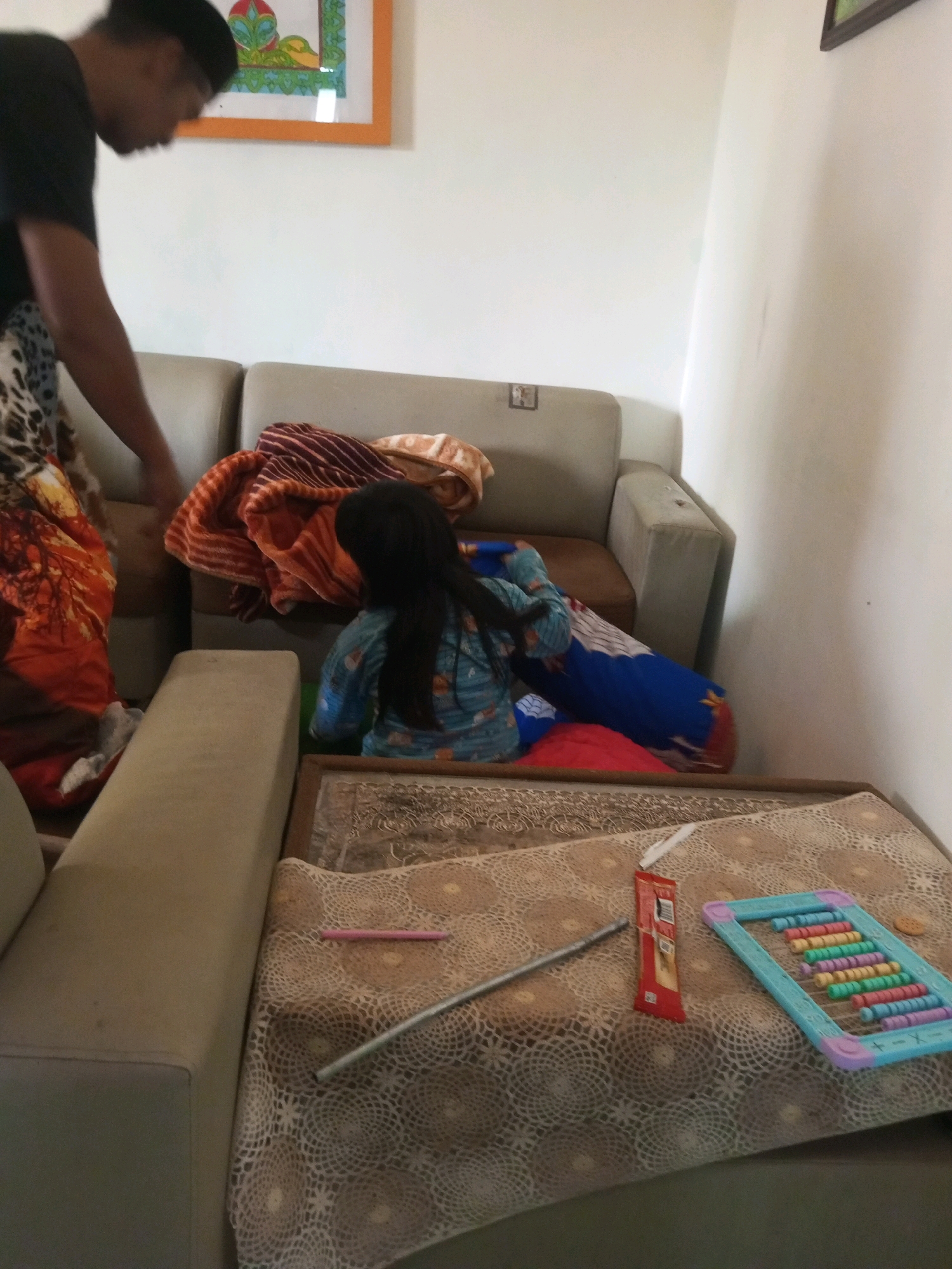 An adult male, partially visible, wearing a black t-shirt, stands near a couch in a room. A young girl, wearing a blue and patterned outfit, is seated on the couch, partially obscured by blankets and pillows. The couch is covered with various blankets and cushions. In front of the couch, on a patterned surface, are several objects including a small abacus with colorful beads, a red candy wrapper, and a long silver rod-like object, possibly a pencil or pen. A framed artwork hangs on the wall in the background. The room appears to be a living area, with neutral-colored walls and furniture. The overall lighting suggests it is daytime.