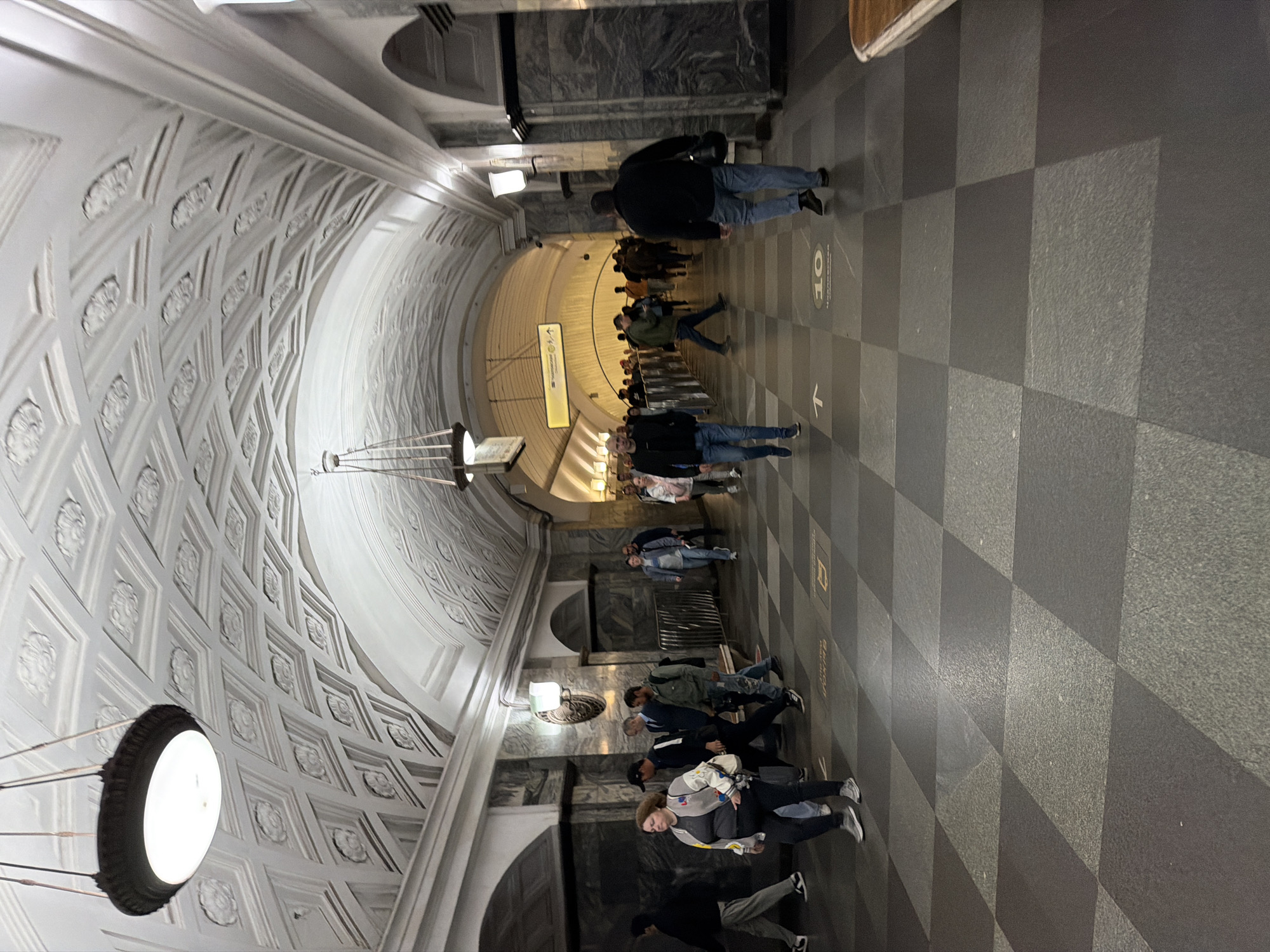 A wide shot captures the interior of a subway station in an unknown city. The vaulted ceiling features ornate detailing and is illuminated by hanging light fixtures. The floor is a checkered pattern of dark and light grey tiles. People are seen walking in both directions along the station platform. A sign with Cyrillic text and an arrow is visible in the background. The walls are lined with dark marble.