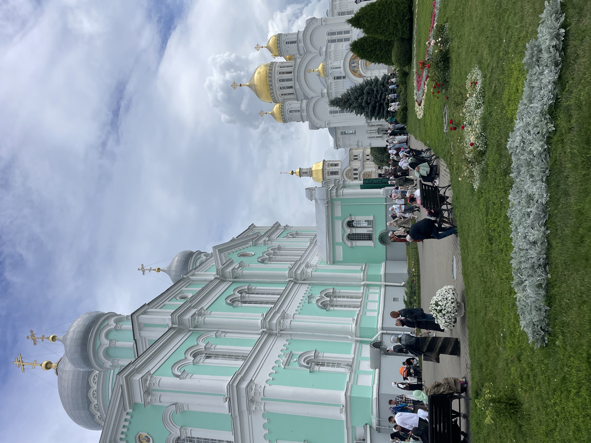 In Diveyevo, Russia, a group of people are gathered on a cloudy day in front of a turquoise and white church with silver domes. To the right, a large white church with golden domes is visible. The people are dressed in modest clothing, many with headscarves, and some are sitting on benches. A pathway is visible between the churches, lined with green grass and flower beds. Several dark green evergreen trees are planted in front of the white church.