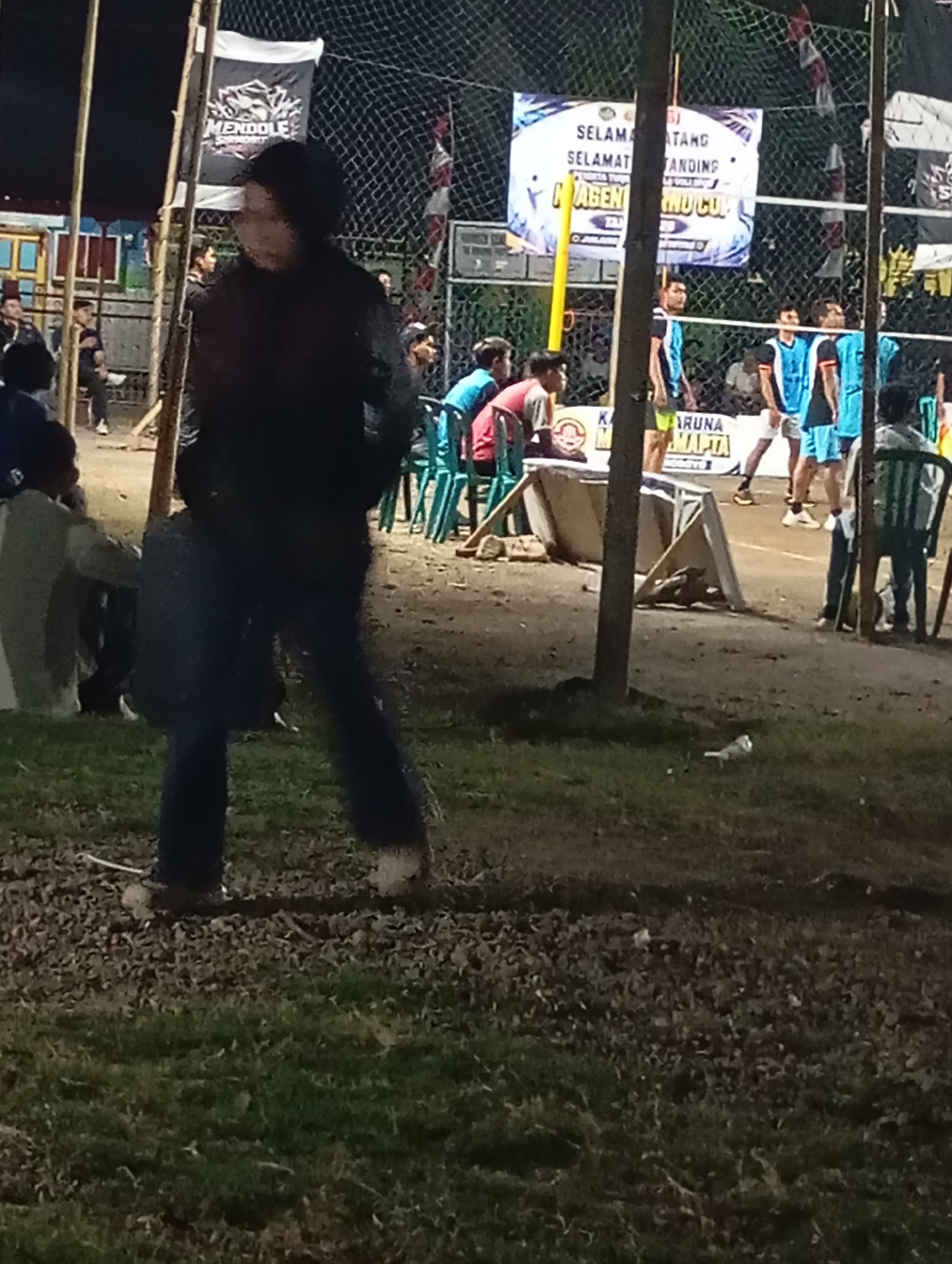The image captures an outdoor evening scene, likely at a sports venue, possibly a volleyball court, given the net and the players in athletic attire. A banner in the background reads "SELAMAT DATANG SELAMAT BERTANDING" and "MAGENTINO CUP". The setting appears to be a community event or tournament.
In the foreground, a person wearing a dark jacket and dark pants is walking, their face mostly obscured by shadow and blur. Their posture suggests movement. To the left, partially visible, are other individuals sitting or standing, also in casual attire. In the mid-ground, several young men in blue and white athletic jerseys are gathered on a sandy surface, some appearing to be players. Green plastic chairs are set up on the sidelines. The ground is a mixture of grass and dirt, with fallen leaves scattered around. The overall lighting suggests it's nighttime, with artificial lights illuminating the area. The presence of the banner and the organized setup indicates a public event. The location is identified as Pacitan, Indonesia, implying a local community gathering in that region.