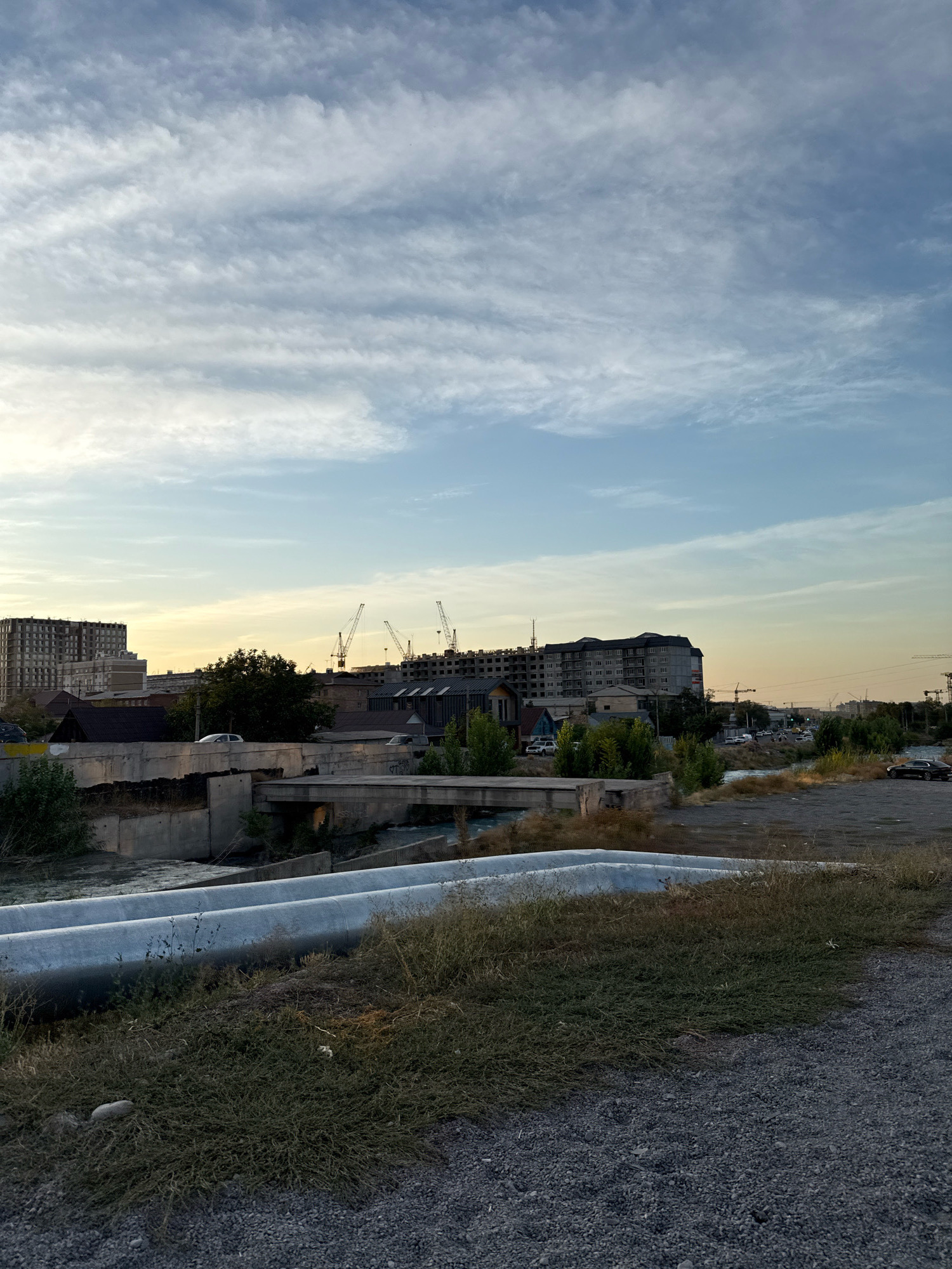 The image depicts an urban landscape in Bishkek, Kyrgyzstan, possibly during the late afternoon or early evening, as indicated by the warm light and soft shadows. In the foreground, two large, light-colored pipes run horizontally across the frame, partially obscured by dry grass and gravel. Beyond the pipes, a river or canal flows, crossed by a concrete bridge. The middle ground features several buildings, including a multi-story residential building under construction and other structures that appear to be a mix of older and newer architecture. Construction cranes are visible above some of the buildings, suggesting ongoing development. Further in the distance, more buildings and trees line the horizon, with a road and a few cars visible. The sky is a light blue, with scattered, wispy white clouds, and a soft yellowish glow near the horizon, suggesting the setting sun. The overall scene portrays a functional, developing urban environment.