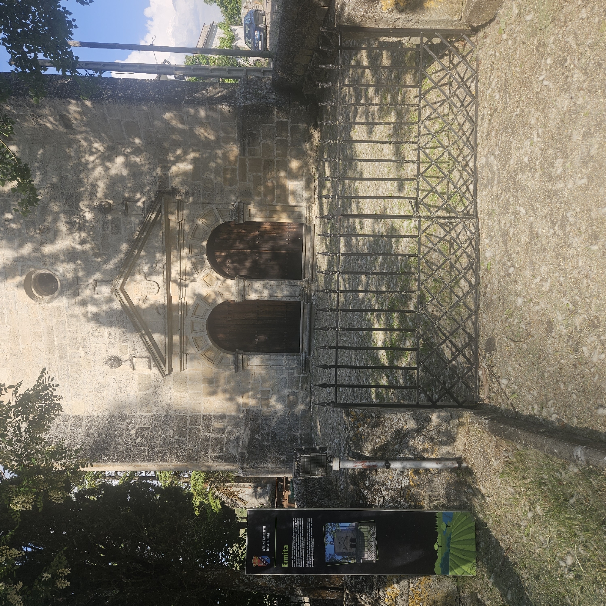 Exterior view of the Ermita de Santa Quiteria in Algora, Spain. The facade of the stone building has two arched doorways and a small circular window. A black metal fence borders the building. A sign with the city crest, "Ayuntamiento de Algora" is posted. The sign, which describes the history of the Ermita, has a black background with green graphics. Trees and vegetation are visible around the building.