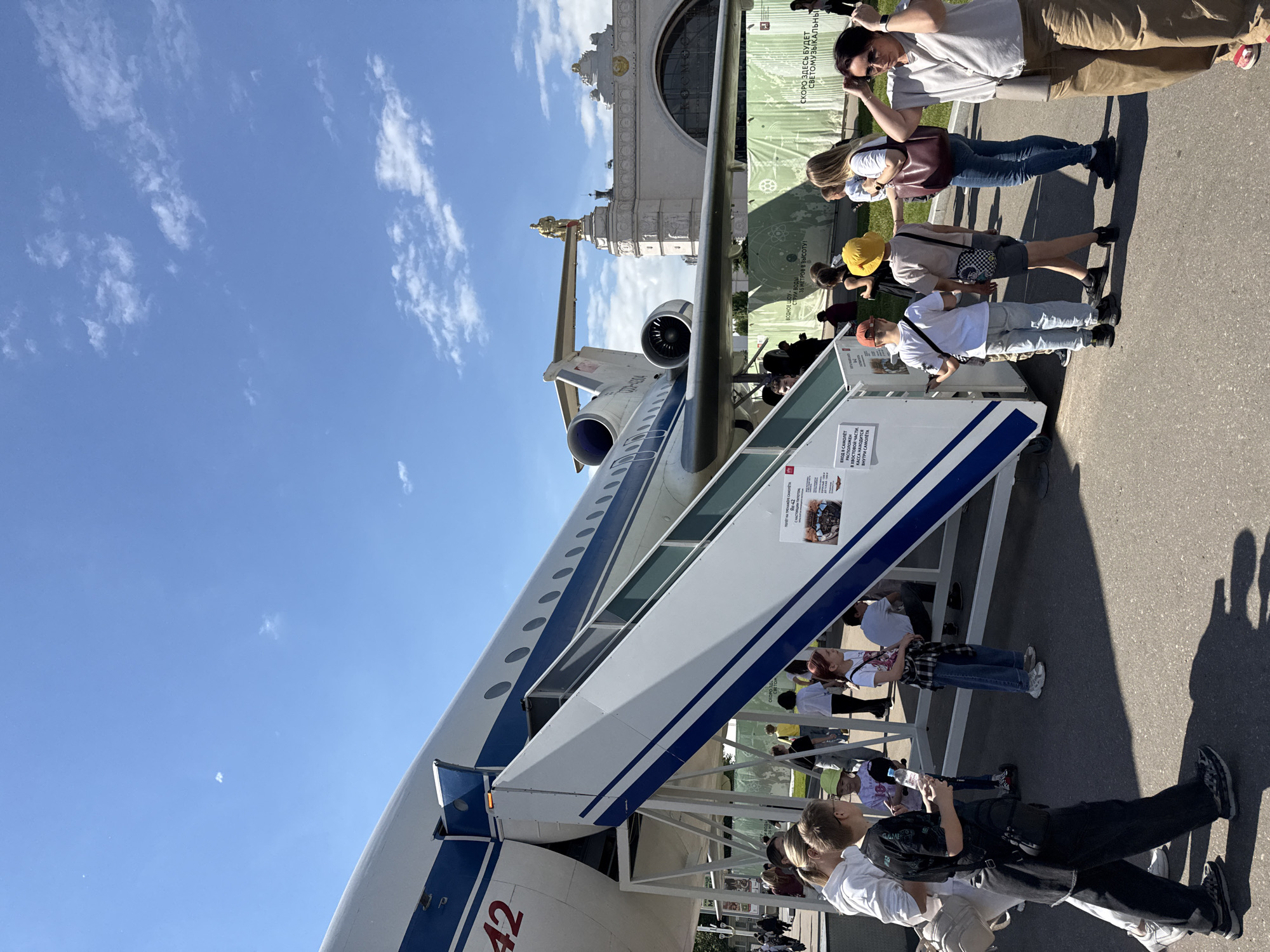 This media file captures an outdoor exhibition scene on a clear, sunny day, likely in Khimki, Russia, featuring a parked aircraft and numerous visitors.
The central subject is a large white turbofan airplane, identified as a Yak-42, with distinct blue stripes along its fuselage. Boarding stairs are extended to its forward left entry door, indicating it's accessible to the public. The tail section of the aircraft is partially visible, showing the number "42".
A diverse crowd of people, casually dressed, is gathered around the aircraft and its stairs. Many appear to be queuing to enter the plane, while others stand nearby, observing or taking photos with their phones. Strong shadows cast on the ground suggest it is mid-day. The sky is bright blue with scattered white, wispy clouds.
In the background, a grand, ornate building with classical architectural elements and a golden statue on its pinnacle stands prominently. A large green banner or wall is situated behind the aircraft and people, featuring text and graphics.
Visible text on a sign near the aircraft stairs confirms it is a "Yak-42 aircraft" (ЯК-42). Other text instructs, "Entrance to the aircraft is located in the tail section" (ВХОД В САМОЛЕТ РАСПОЛОЖЕН В ХВОСТОВОЙ ЧАСТИ) and "Ticket office is inside the aircraft" (КАССА НАХОДИТСЯ ВНУТРИ САМОЛЕТА), indicating it's an interactive exhibit. The green banner advertises other attractions: "Water show" (ВОДНОЕ ШОУ-), "Water jets 35 meters high!" (СТРУИ ВОДЫ 35 МЕТРОВ В ВЫСОТУ!), and "Soon there will be a light and music show" (СКОРО ЗДЕСЬ БУДЕТ СВЕТОМУЗЫКАЛЬНЫ). The word "Космо" (Cosmo/Space) is also visible on this banner.
The scene depicts a lively public event or exhibition, with people exploring and interacting with a static aircraft display amidst other advertised attractions.