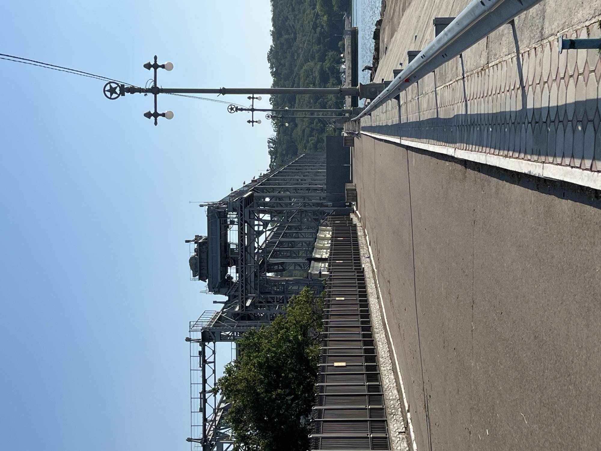The image depicts an industrial scene with a large metal structure, possibly a conveyor or crane, dominating the left side. This structure is a complex assembly of beams, platforms, and machinery, suggesting a place of industrial activity. To the right of this structure is a paved walkway or road, bordered by a decorative stone pattern. Beyond the walkway, a metal railing runs along the edge of what appears to be a pier or embankment, with the calm blue water of a harbor or sea visible in the background. A lush green hillside covered in trees rises beyond the water, creating a natural contrast to the industrial elements. The sky is a clear, bright blue, indicating a sunny day. The presence of streetlights on tall poles suggests this is a developed area, possibly a port or industrial zone. No people are visible in the frame. The overall impression is one of a functional, outdoor industrial setting on a clear day.