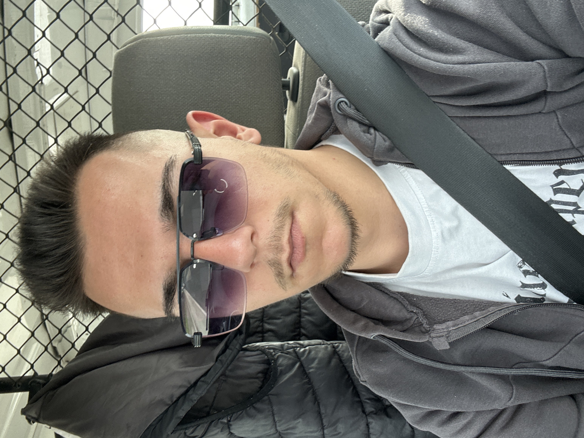 A man is seated in a vehicle, wearing sunglasses, a grey hoodie, a white t-shirt, and a black quilted jacket. He is wearing a seatbelt. A fence is visible through the window behind him. The location is unknown.