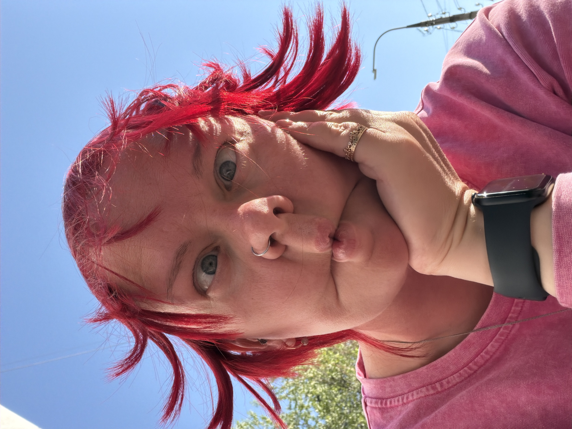 A low-angle selfie of a person with dyed red hair. The person is wearing a pink shirt and a black smartwatch on their left wrist. The person has a nose ring and is making a pouting facial expression while holding their right hand against their cheek. The background includes a bright blue sky, a utility pole with wires, and some trees. The exact location within the city of Chaikovsky, Russia is not discernible from the image.