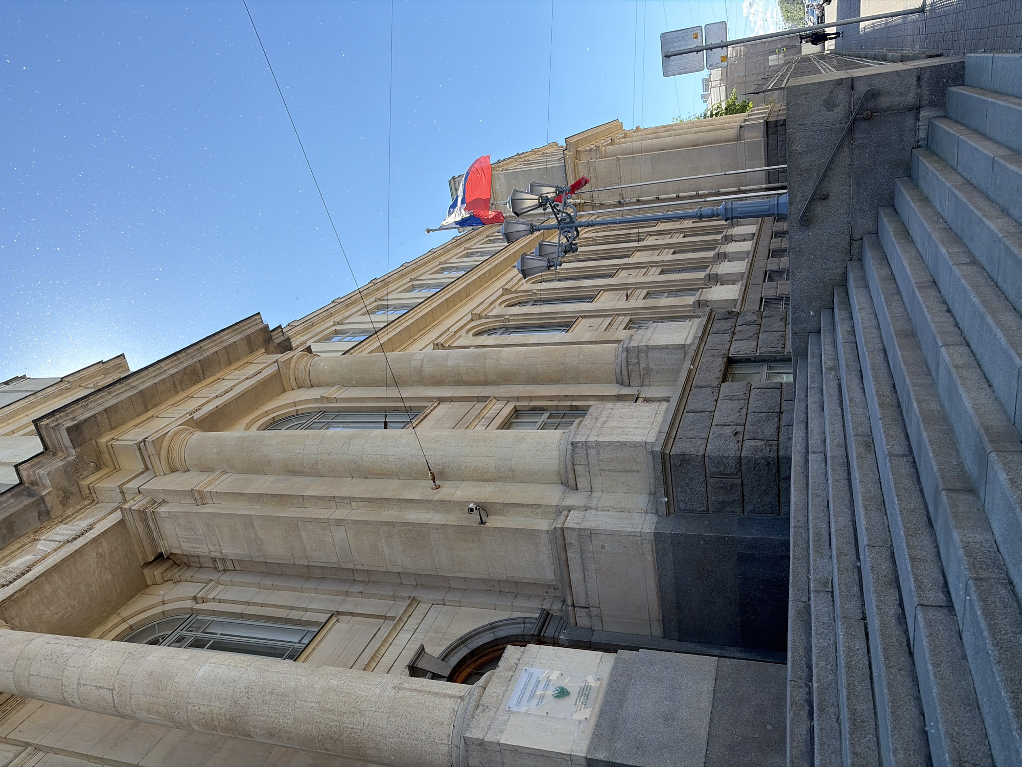 The image depicts the exterior of a large, classical-style building, likely an educational institution or government building, situated in Saint Petersburg, Russia. The architecture features prominent stone columns, arched windows, and a stone facade. A wide, stone staircase leads up to the building's entrance.
In the foreground, the stone steps are well-lit by the sun, with some granular debris visible on their surface. To the left of the steps, a portion of the building's lower section is visible, clad in dark granite blocks. A sign is affixed to the wall near the entrance.
Further back, a lamppost with multiple lights stands near the building. Two flags are visible, with the larger one, a Russian tricolor flag, fluttering in the breeze. Another, smaller flag is partially obscured. The sky above is a clear, bright blue, with numerous small white specks, possibly dust or pollen, suspended in the air and illuminated by the sun.
To the right of the building, a sidewalk is visible, lined with a low stone wall and a metal fence. A traffic sign is mounted on a pole. A single person is walking on the sidewalk in the distance. The street appears to be paved, and some vehicles are present further down. The overall impression is a sunny day in an urban environment with significant architectural heritage.