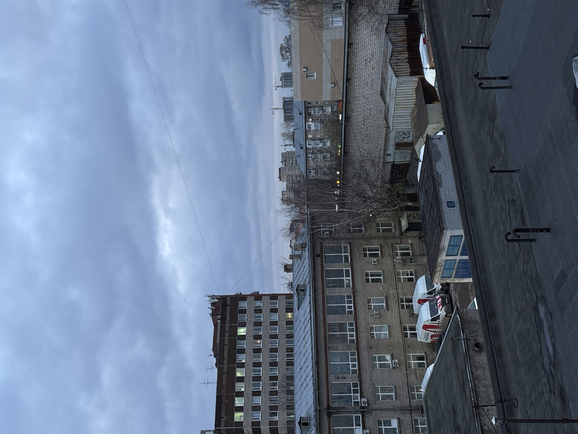 The image shows a cityscape, possibly taken from a high vantage point in Perm, Russia, based on the architectural style of the buildings. The sky is overcast, suggesting a cloudy or potentially rainy day. In the foreground, there is a flat rooftop with metal fixtures, possibly ventilation pipes or antennae mounts. Adjacent to the roof, there are several buildings. A tall, Soviet-era apartment building with a concrete facade and rows of windows is visible, along with shorter brick buildings with numerous windows and air conditioning units. Several parked delivery vans are parked in a courtyard. In the background, the city extends with various residential buildings and construction cranes, hinting at ongoing development. Trees are interspersed among the buildings, many of which are without leaves.