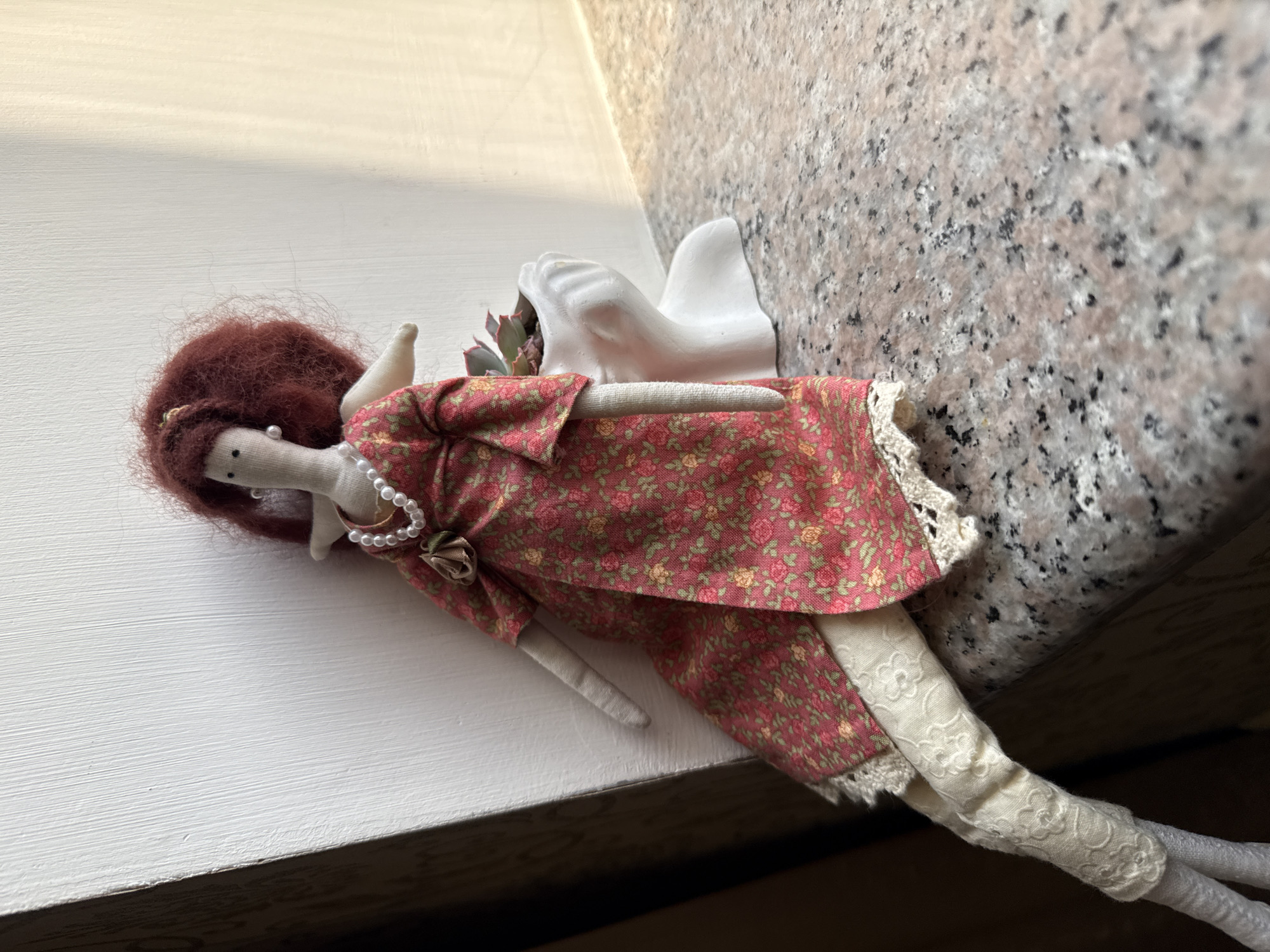 A cloth doll with curly red hair is sitting on a granite countertop, leaning against a white wall. The doll is wearing a floral print dress with lace trim and cream-colored leggings with a floral pattern. A string of pearls is around the doll's neck. To the right of the doll is a white planter in the shape of a face, containing a small succulent plant. The background shows a wall and a dark area to the left, possibly a window frame or furniture. The scene is lit by natural light, casting shadows. The location is not specified within the city of Sevan, Armenia; the context of the query is likely a mistake or misinterpretation of the provided information.