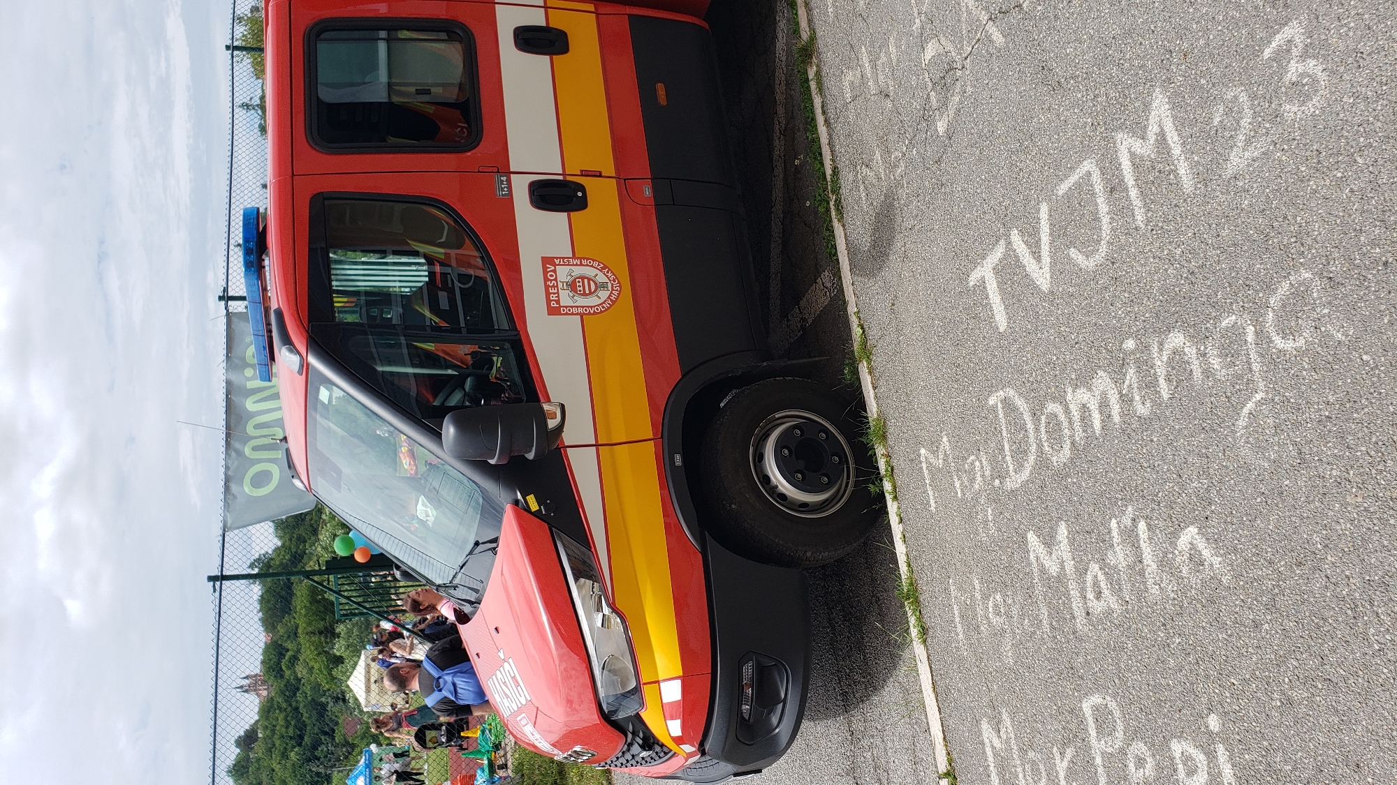 A red and black fire brigade vehicle, identified by the "PREŠOV DOBROVOĽNÝ HASIČSKÝ ZBOR MESTA" (Prešov Volunteer Fire Brigade of the City) emblem on its side, is parked on an asphalt surface. The vehicle, appearing to be an Iveco Daily model, features a white and yellow horizontal stripe running along its side, reflective red and white markings on the front bumper, and blue emergency lights mounted on the roof. A "1+1+4" crew capacity indicator is visible near the rear door.
In the mid-ground, a chain-link fence separates the vehicle from an outdoor gathering. Several individuals are visible around white tents, and green and orange balloons are present. A green banner with the partial text "omnn" (likely "Omnia") is attached to the fence. The background reveals a tree-covered hill, topped by a historical structure with twin spires, consistent with architecture found in Prešov. The sky is overcast.
The asphalt pavement in the foreground features multiple white chalk inscriptions, including legible words such as "Dominik," "Maja," and "TVJM 23." The overall scene suggests a public event or gathering in Prešov, Slovakia, likely attended by the local volunteer fire brigade.
