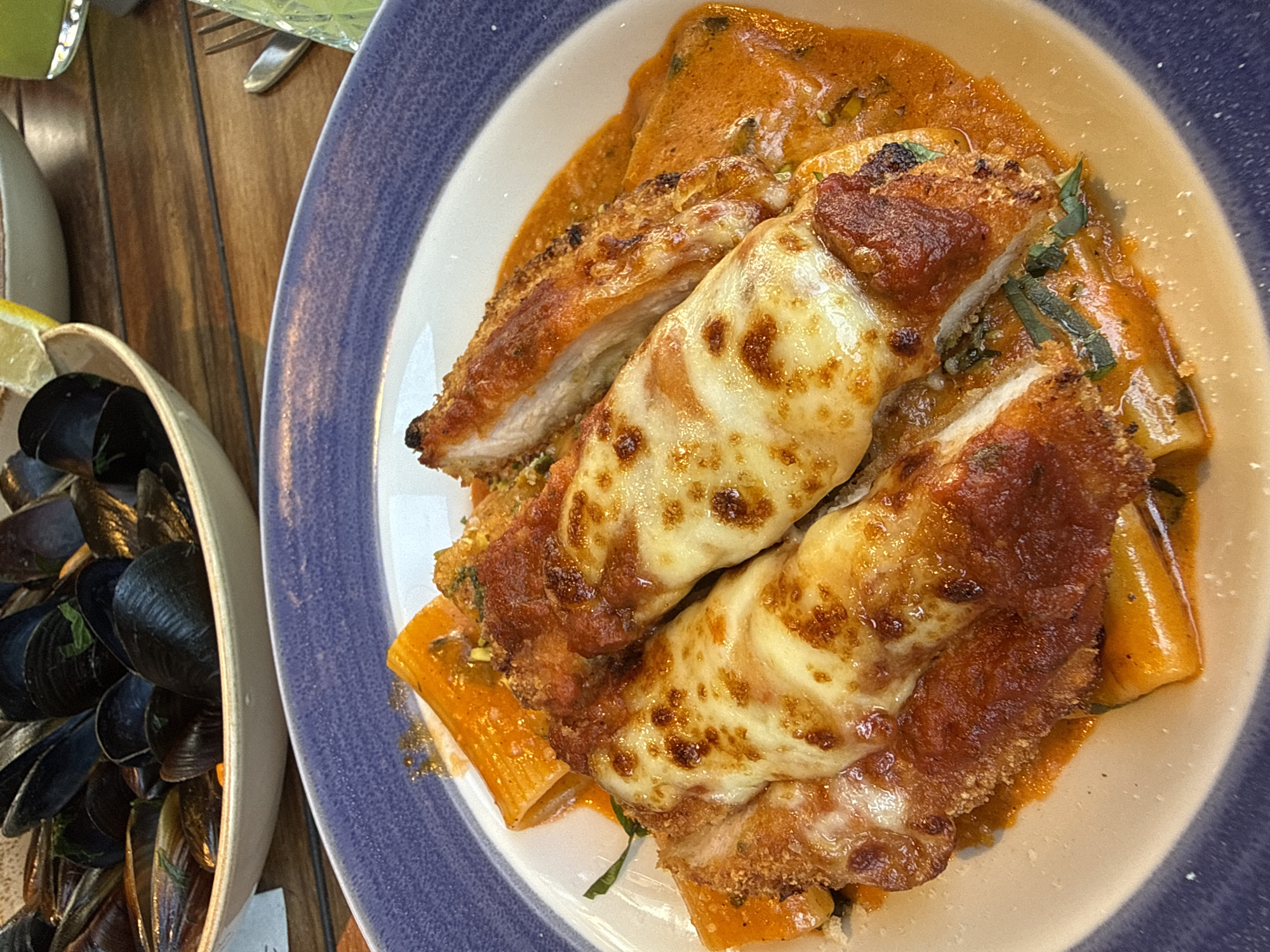 A close-up, overhead shot shows a plate of rigatoni pasta topped with chicken parmesan and a rich, orange sauce. The chicken is breaded and fried, then covered in melted, golden-brown mozzarella cheese and a tomato-based sauce. Some green herbs are visible sprinkled on top. To the left of the pasta dish, a bowl of steamed mussels in their dark shells is visible, with a slice of lemon on the rim of the bowl. The food is presented on a wooden table, with the edge of a blue and white patterned plate visible in the foreground. The overall scene suggests a meal at a restaurant. No people are visible. The lighting appears to be natural, possibly from a window, and the time of day is unclear. There is no visible text.