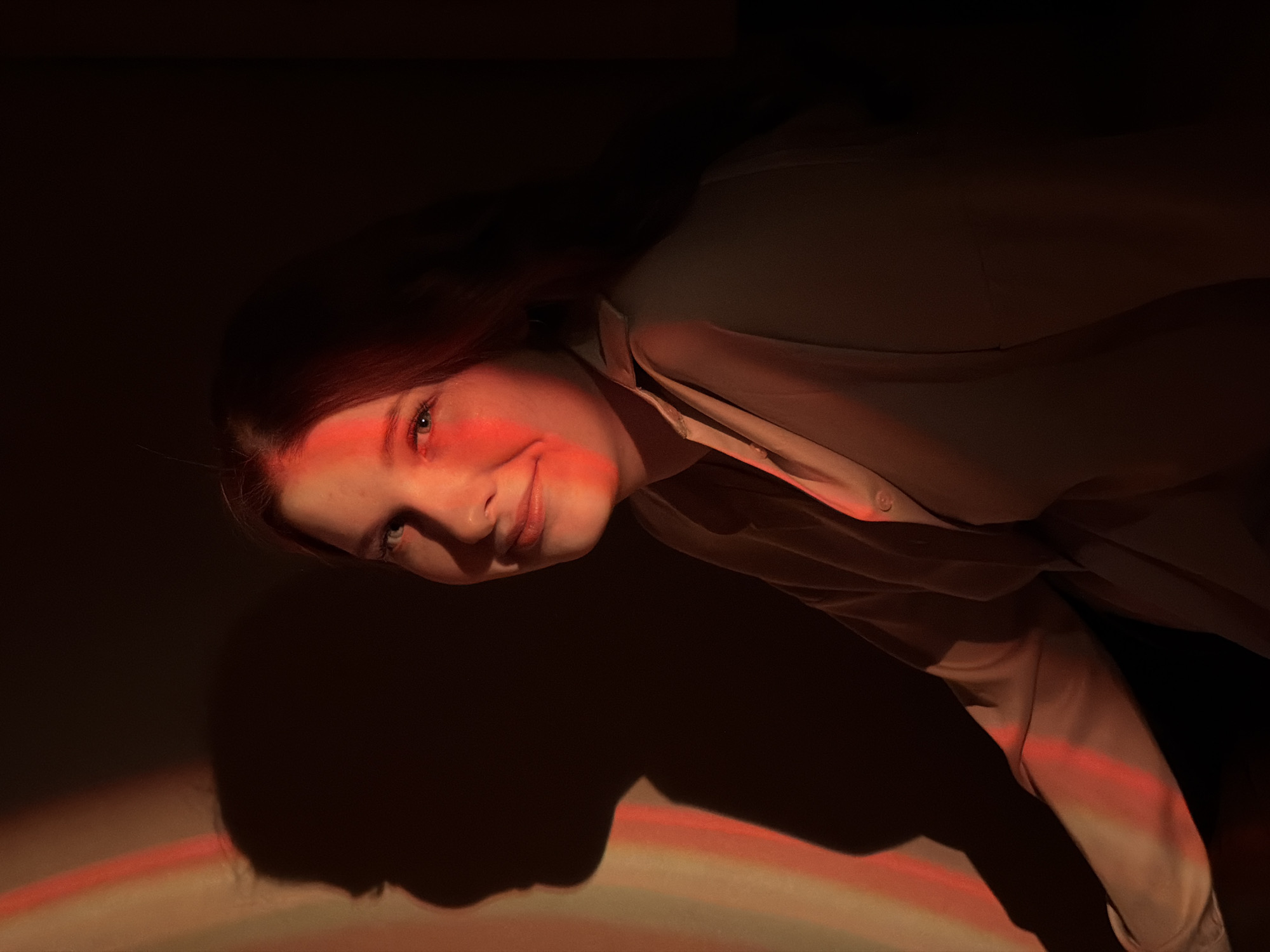 The image captures a young woman in Chaikovsky, Russia, within a dimly lit indoor setting, dramatically illuminated by a red-orange light. The photograph is taken from a slightly elevated and tilted perspective, showing the woman looking upwards and towards the camera with a subtle smile or smirk. Her fair skin, light-colored eyes, and portions of her reddish-brown hair are bathed in the warm, intense glow, creating striking highlights and shadows on her face.
She is wearing a light-colored, possibly beige or off-white, collared shirt or blouse, which also reflects the ambient red light. The background is predominantly dark, drawing focus to her face and the unique lighting. Below and to the left of her, a distinct shadow of her head and upper body is cast on a surface, also partially caught by the red light, creating an interesting effect with bands of color within the shadow, resembling a faint rainbow.
The scene suggests an artistic or studio portrait, utilizing controlled lighting to evoke a specific mood—possibly contemplative, playful, or intense. No specific location cues other than the indoor setting are visible, and the time of day appears to be nighttime or an intentionally darkened environment. No visible text is present in the image.