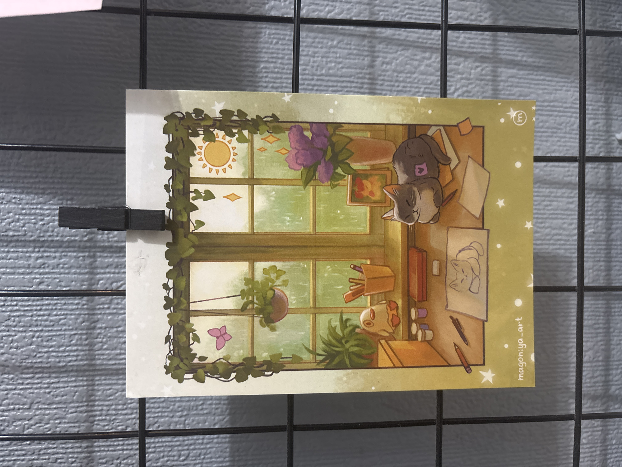 A print is clipped to a wire grid. The print depicts an illustration of a windowsill scene. A window with a grid pattern looks out onto a green, rainy landscape with a sun visible in the upper right corner. Green vines with leaves frame the top of the print. On the windowsill, there is a potted plant, a vase with purple flowers, a framed picture, and various art supplies including pencils, pens, and paint pots. A small, white, duck-like figurine with orange feet sits on the windowsill. A grey cat with a pink heart on its back is curled up on a stack of papers. In the foreground, there are more papers with a sketch of a cat on one of them, and two pencils. The bottom of the print has the text "magon:ya_art" and a small circular logo with the letter 'm' inside a circle. The print is lit from the right, casting a slight shadow to the left. The background is a textured grey surface. This scene is not located in Sinyavino, Russia.
